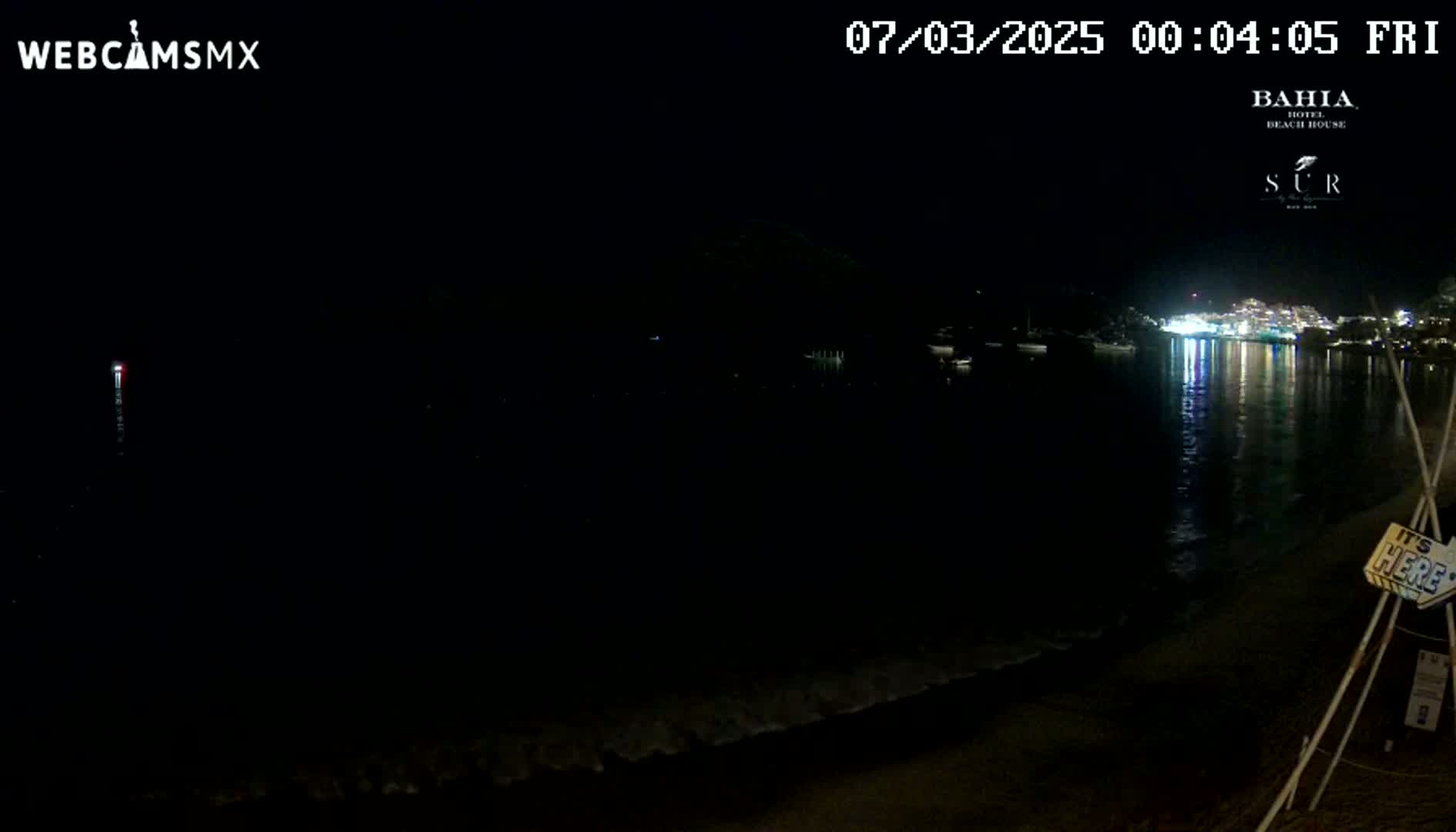 A dark nighttime beach scene shows a calm body of water reflecting the lights of a town across the bay, under a clear sky.