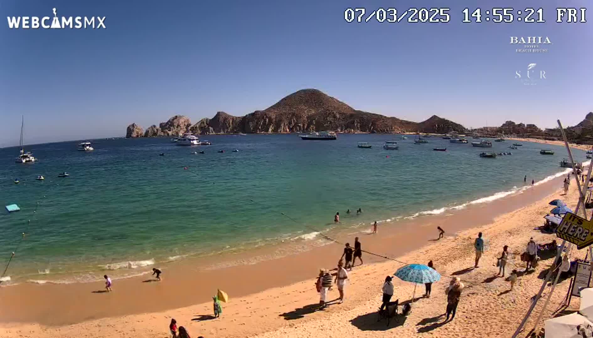 A sandy beach with calm, clear turquoise water and several boats in a bay, under a bright sunny sky, features people relaxing and swimming.