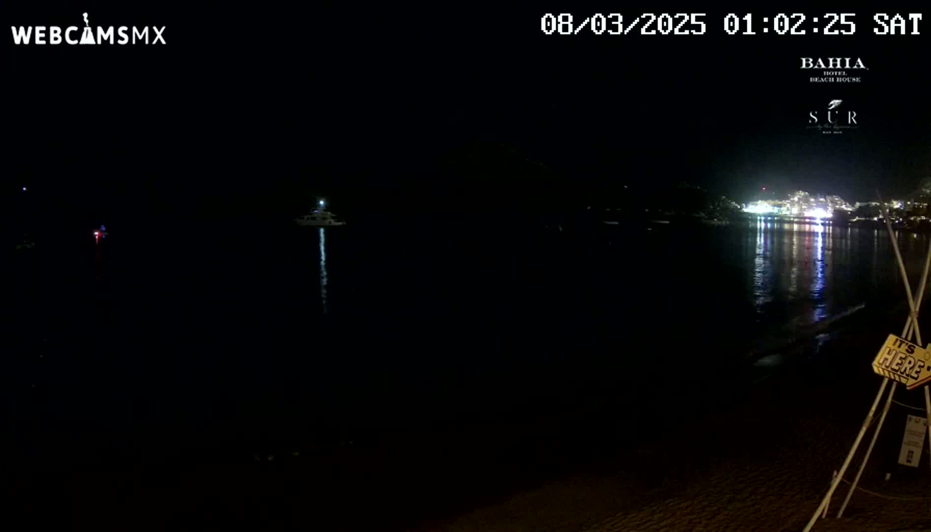 A dark nighttime scene shows a body of water reflecting lights from buildings in the distance, with a few boats visible on the water and a sandy shore in the foreground.