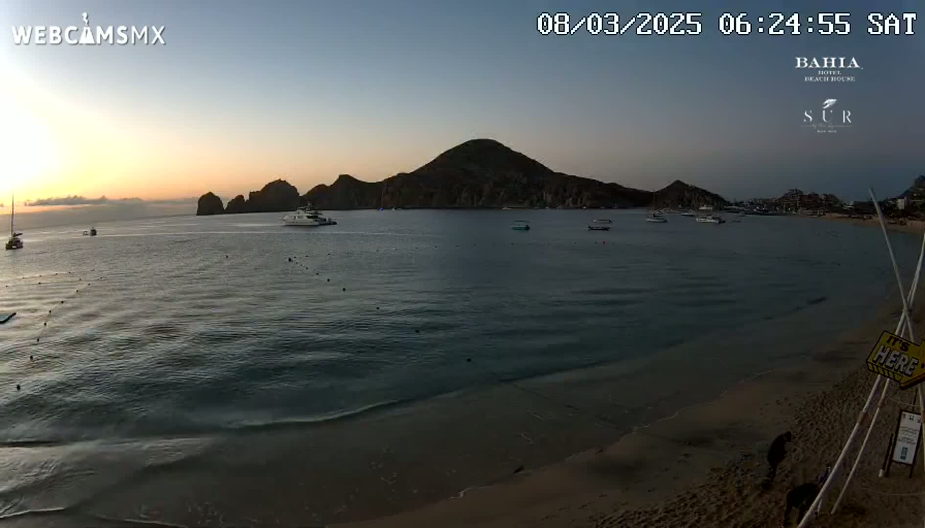 A calm ocean at sunrise with several boats and a sandy beach,  backed by dark, rocky hills.