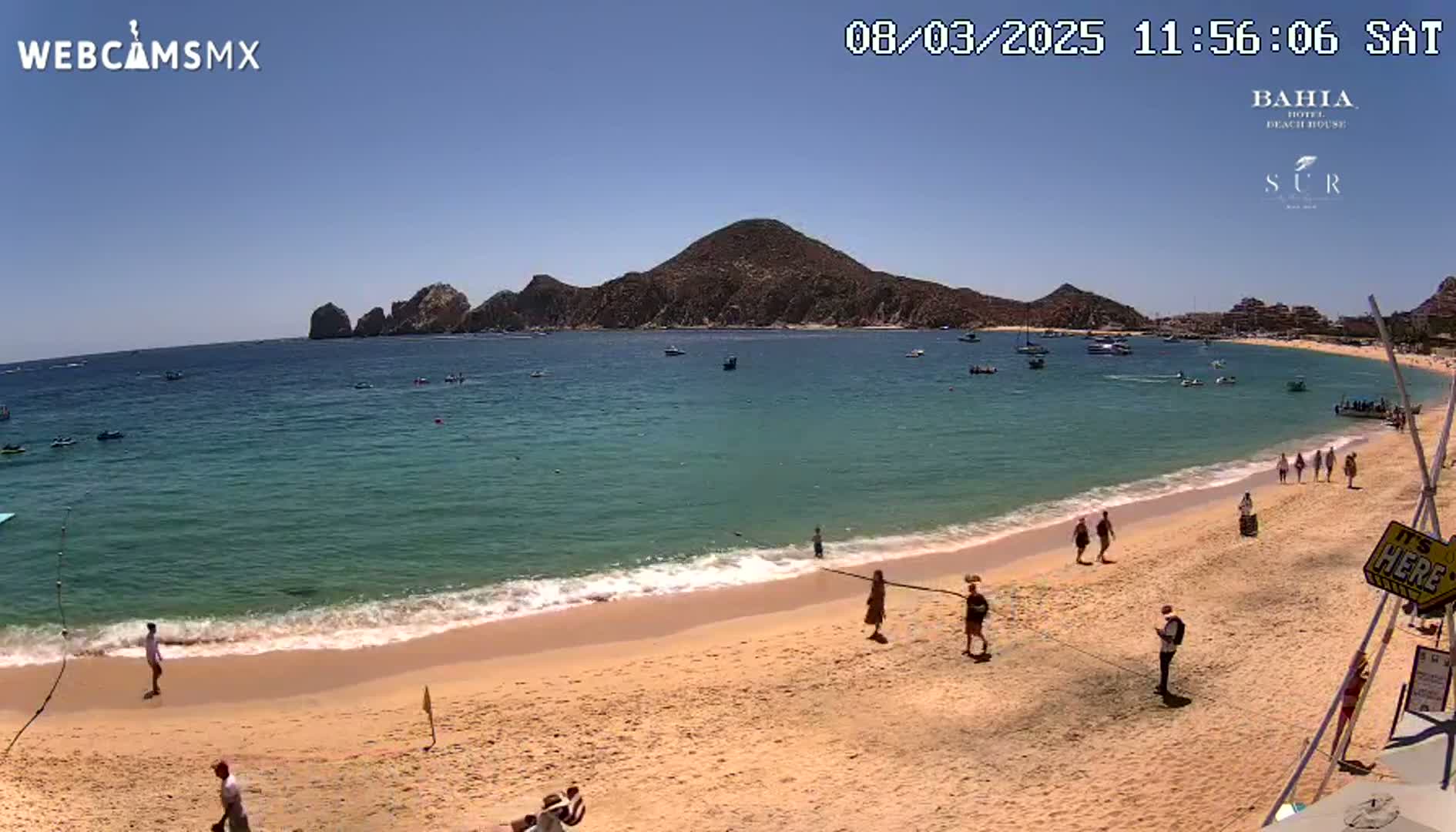 A sandy beach with turquoise water and a few people walking along the shore under a clear, sunny sky, with a large landmass in the background.