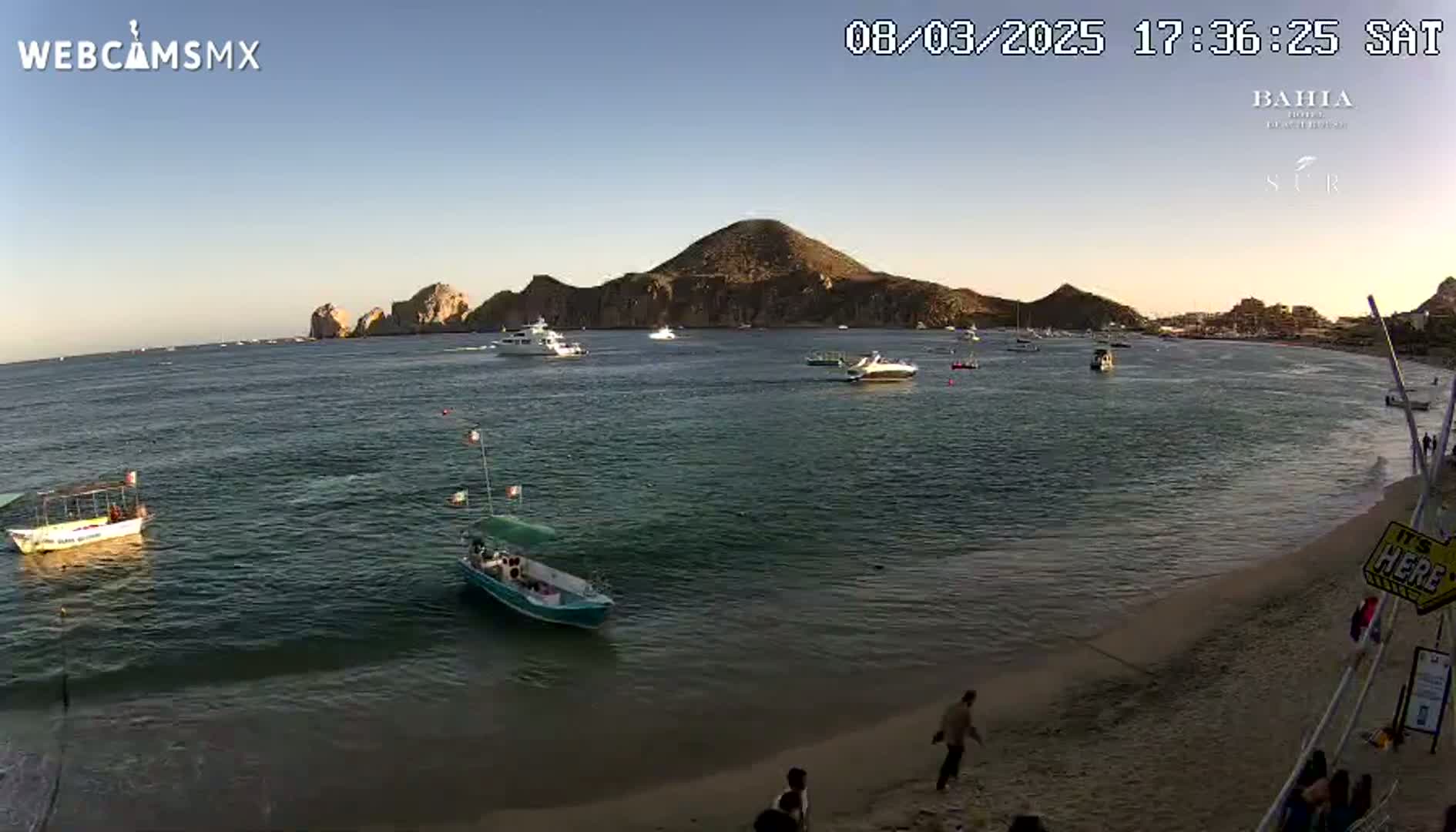 A calm bay with several boats and a sandy beach, under a clear, sunset sky, shows a mountainous peninsula in the background.