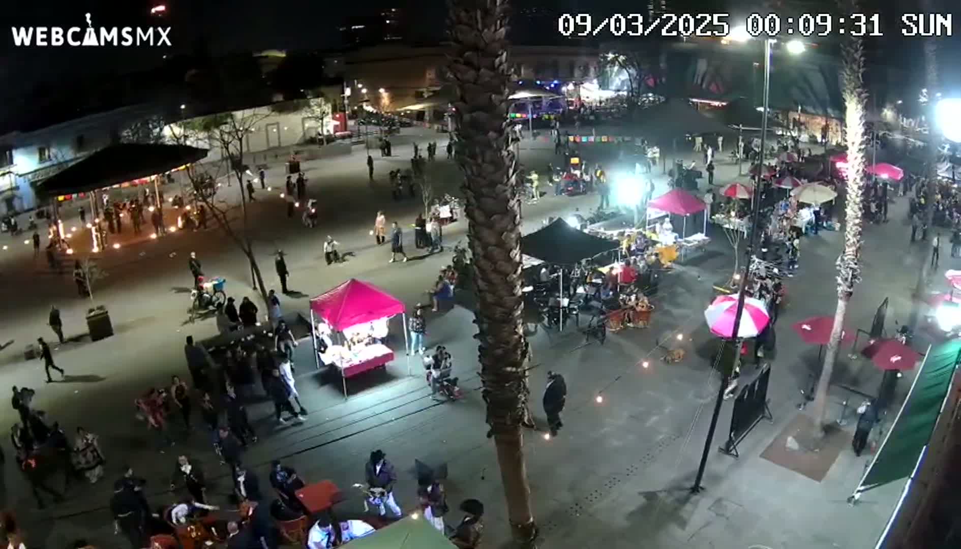 A nighttime aerial view of a bustling plaza with numerous people, food stalls, and market umbrellas under clear skies.