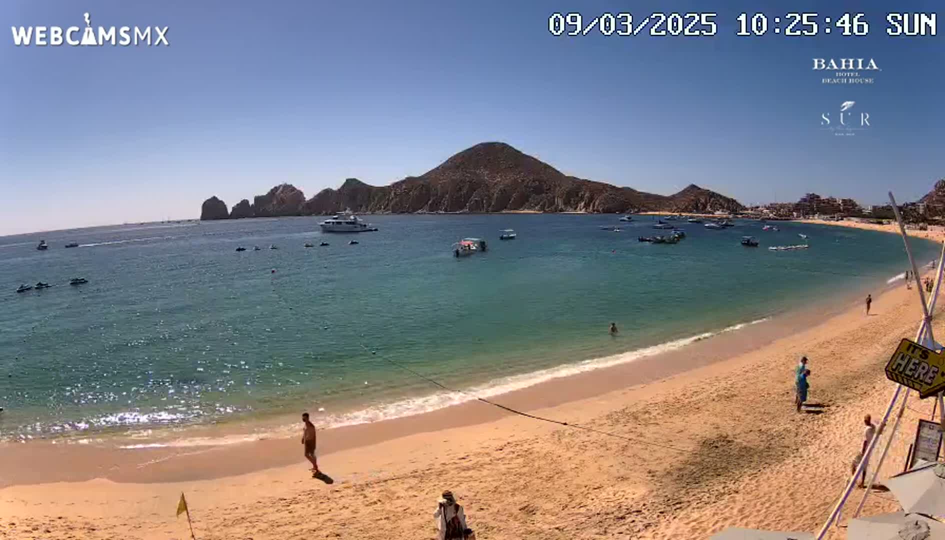 A sandy beach with calm, clear turquoise water, boats in the bay, and a few people on the shore under a sunny sky.