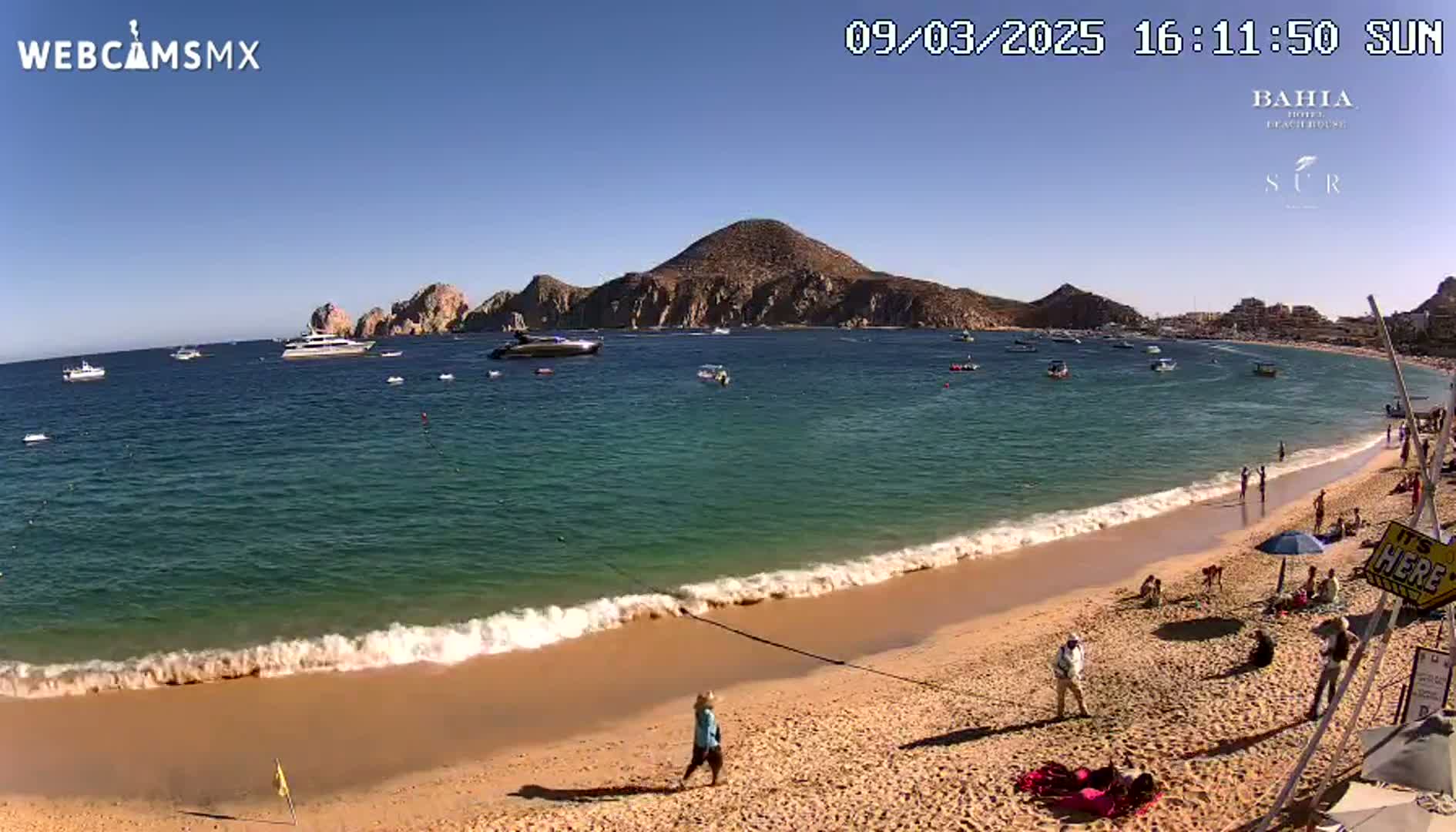 A sandy beach with calm waves laps the shore under a clear blue sky, boats are visible in the turquoise water beyond a rocky peninsula.