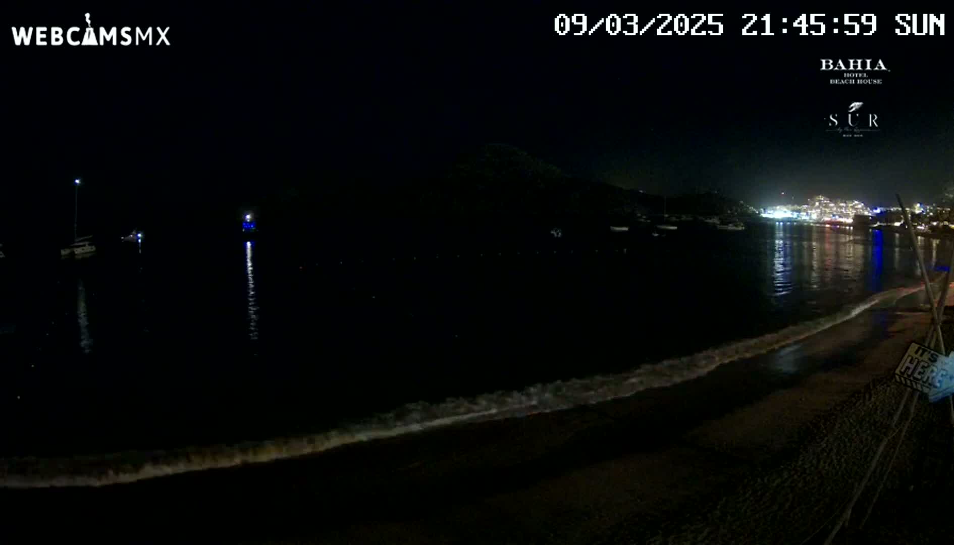A dark, nighttime beach scene shows a calm body of water with boats in the distance, a gently lapping shoreline, and city lights illuminating the opposite side of the bay.