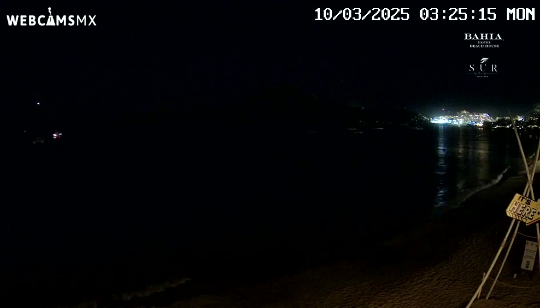 A dark nighttime beach scene shows a calm body of water reflecting the lights of a distant town under a clear sky.