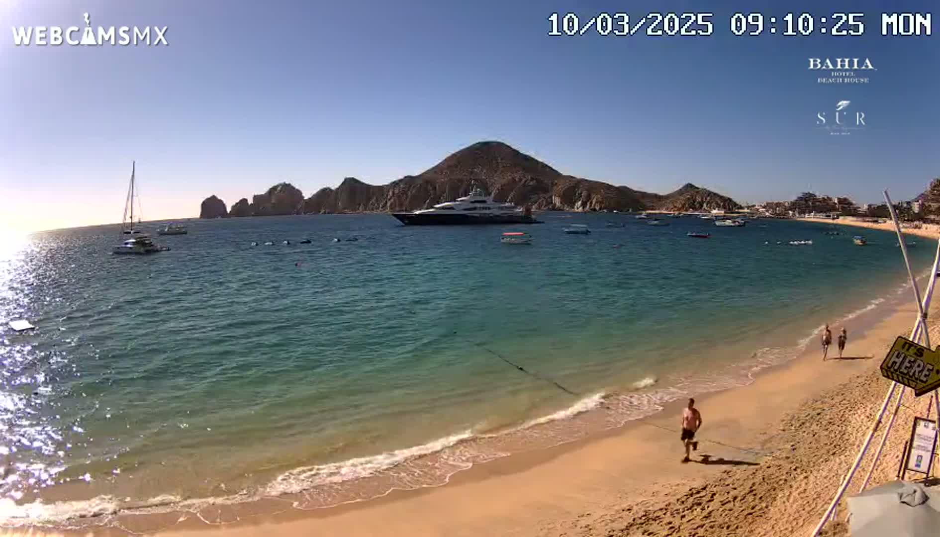 A sandy beach with calm, clear turquoise water laps gently at the shore under a bright sunny sky, with several boats and a large yacht in the bay beyond a rocky peninsula.