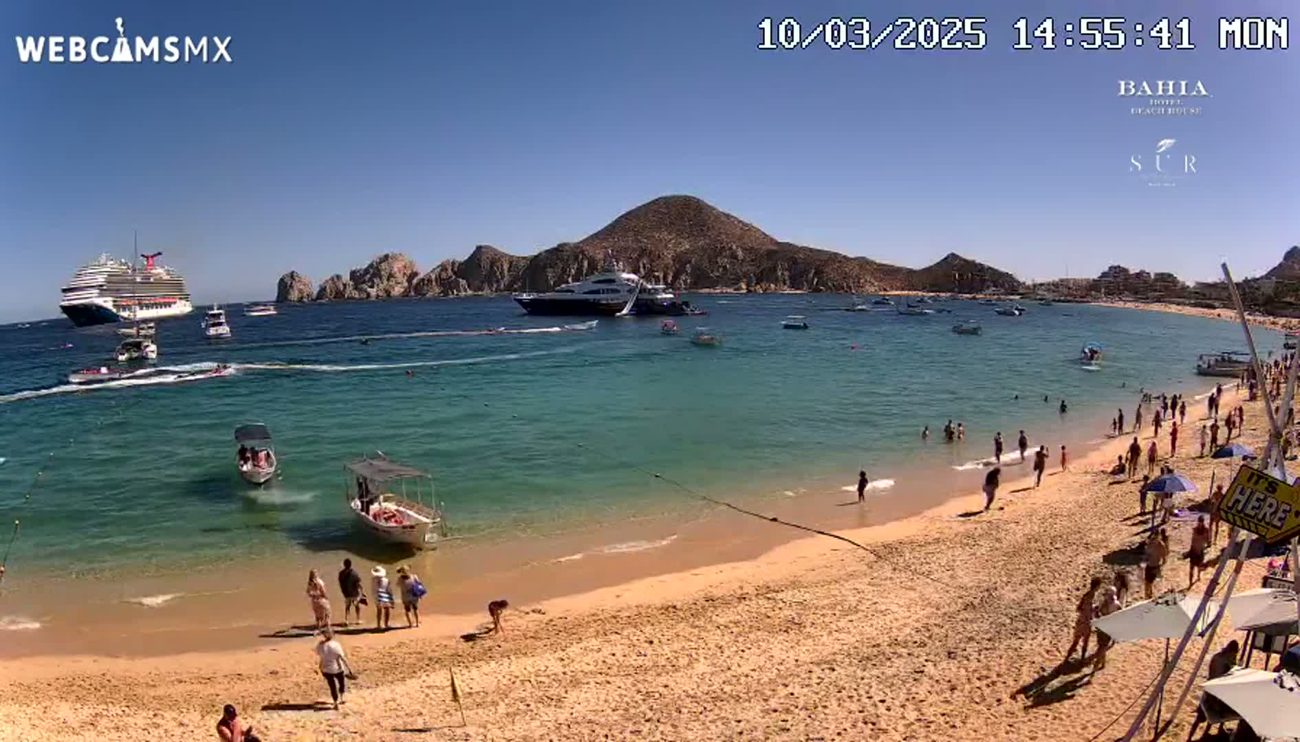 A sandy beach with numerous people, small boats, and a large cruise ship in calm, clear turquoise water under a bright sunny sky, with a mountainous backdrop.