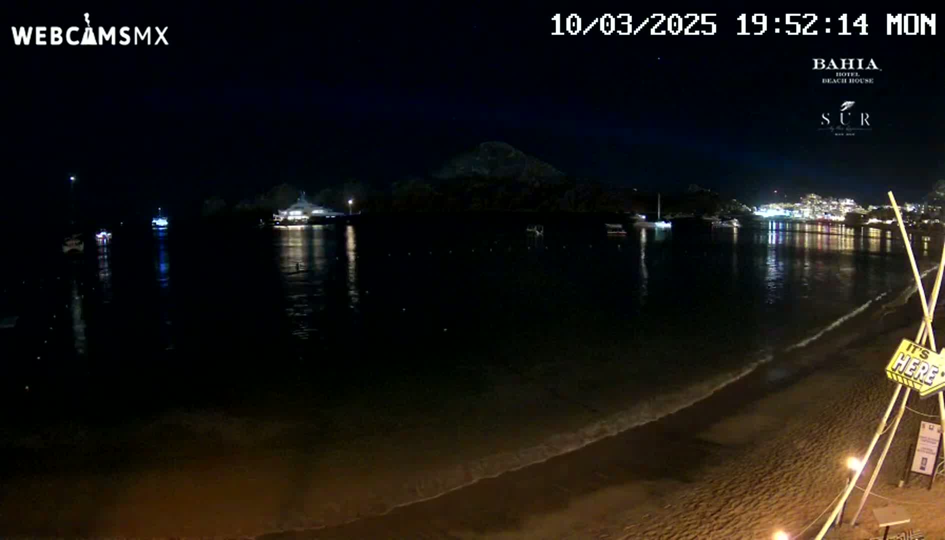 A nighttime beach scene shows calm water reflecting the lights of a town across the bay, under a clear, dark sky.