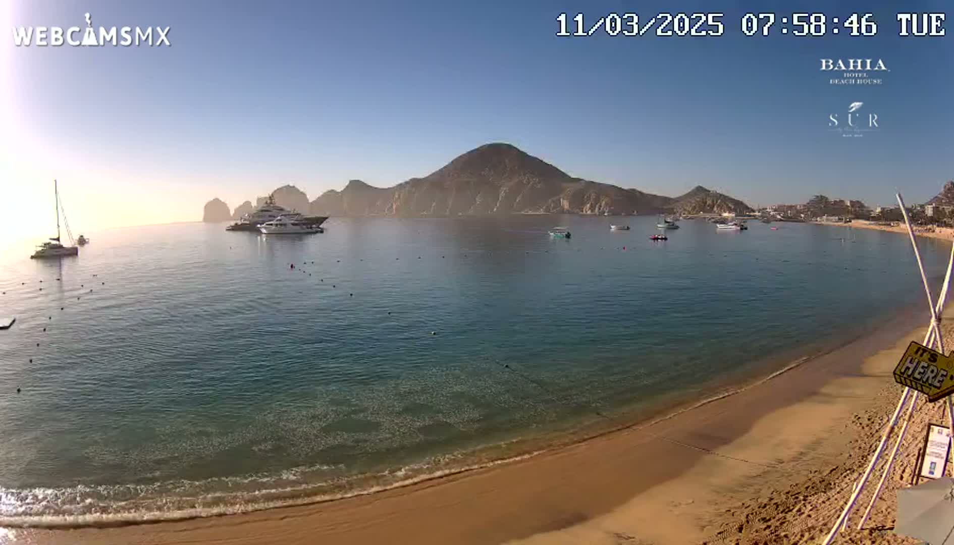 A sandy beach with calm, clear water laps the shore, and several boats are visible in the bay, with mountains in the background under a clear, sunny sky.