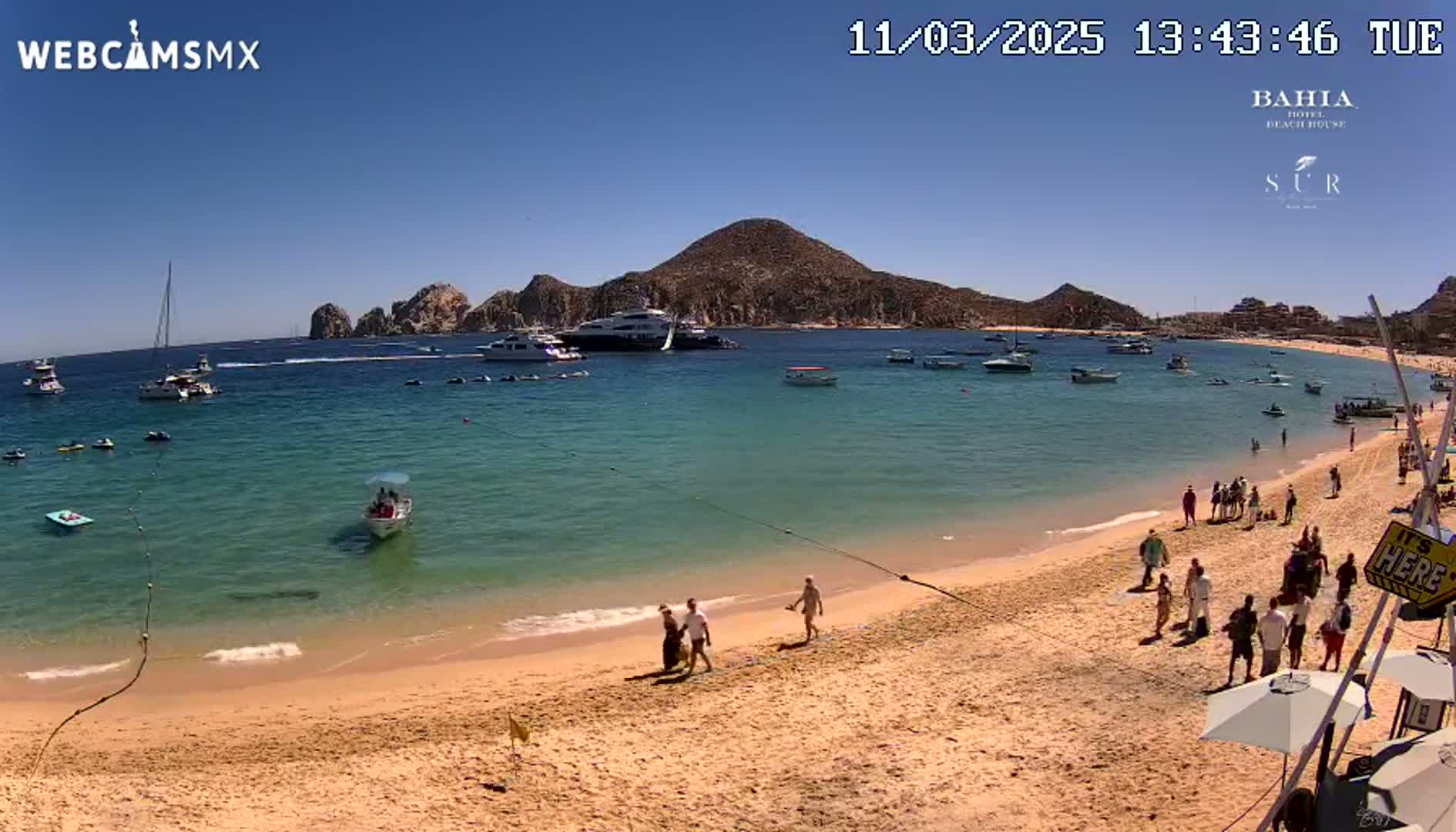 A sandy beach with numerous people and several boats in the clear, blue water under a sunny sky, backed by a rocky hill.