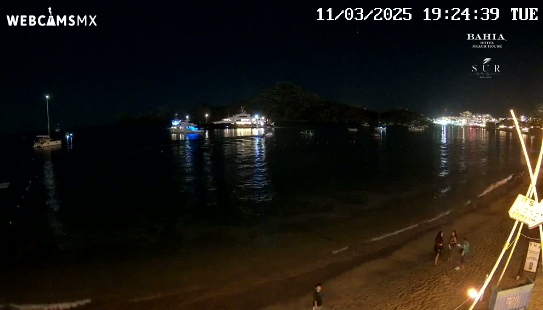 A nighttime beach scene shows several boats in a calm bay, with a few people on a sandy shore under a clear, dark sky.