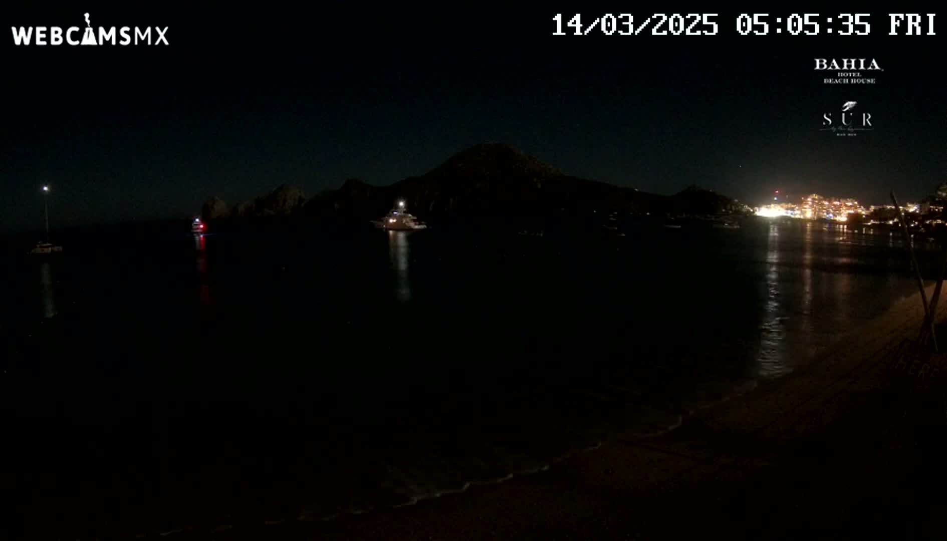 A dark nighttime beach scene shows calm water reflecting lights from a distant town across a bay, with a few boats visible on the water and a hill in the background.