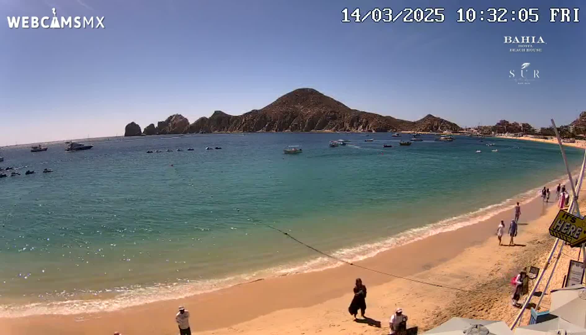 A sunny beach scene shows a calm, turquoise body of water with boats and jet skis, a sandy shore with a few people, and a large, rocky island in the background.
