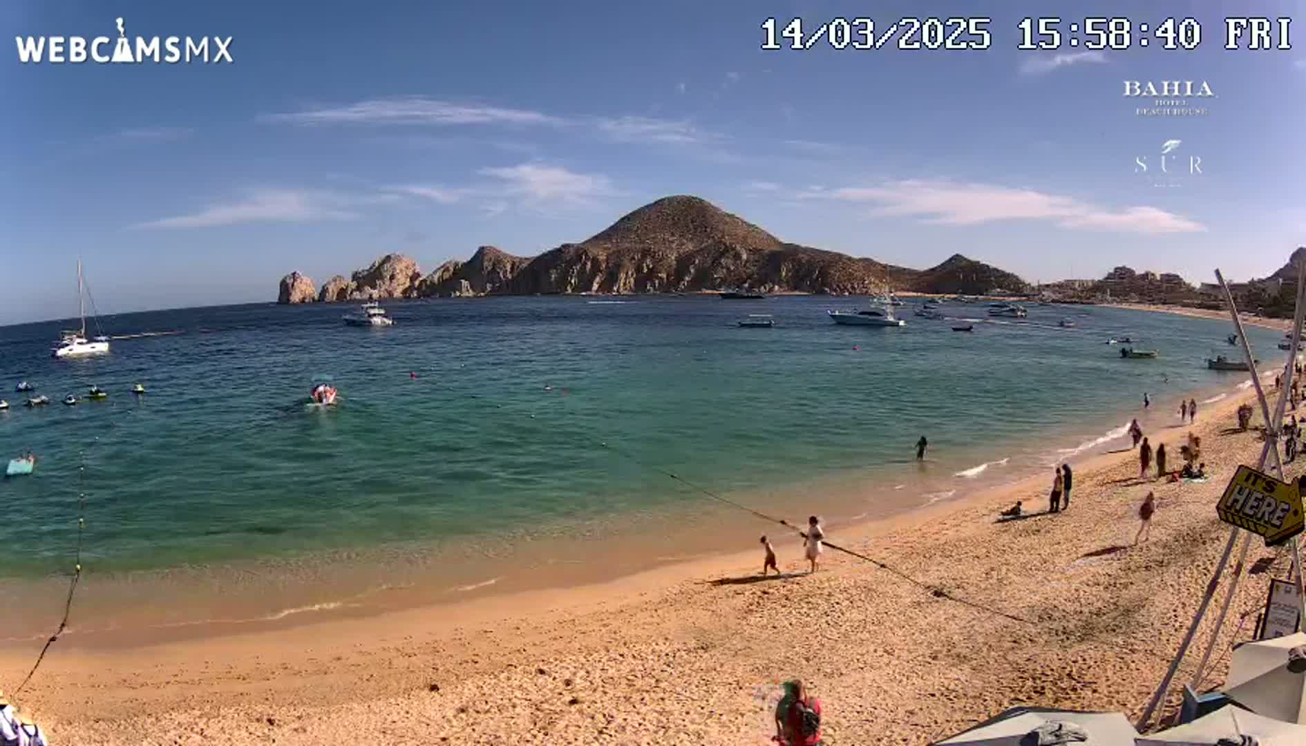A sandy beach with turquoise water and several boats in the bay under a sunny sky, with people swimming and relaxing on the shore.