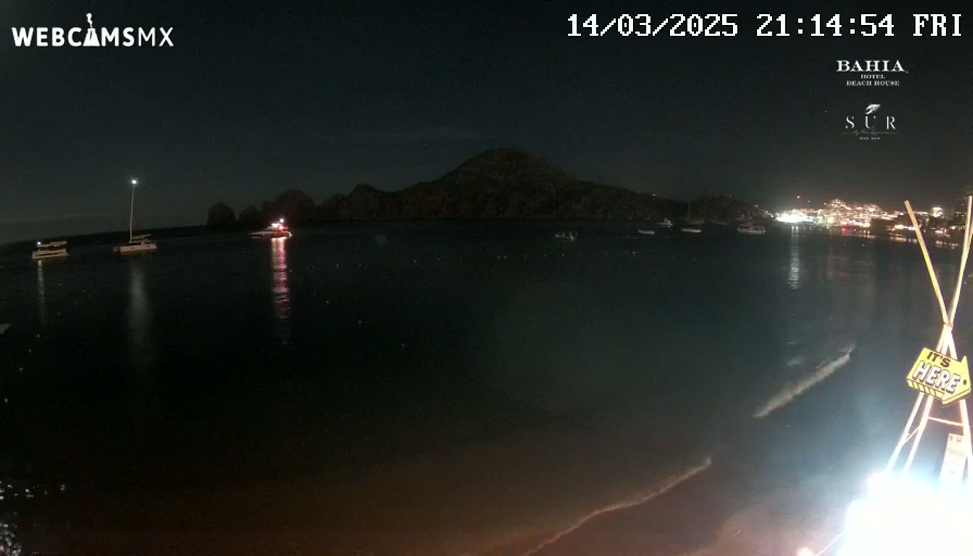 A nighttime view of a calm body of water with several boats, a distant hill, and a brightly lit town in the background.