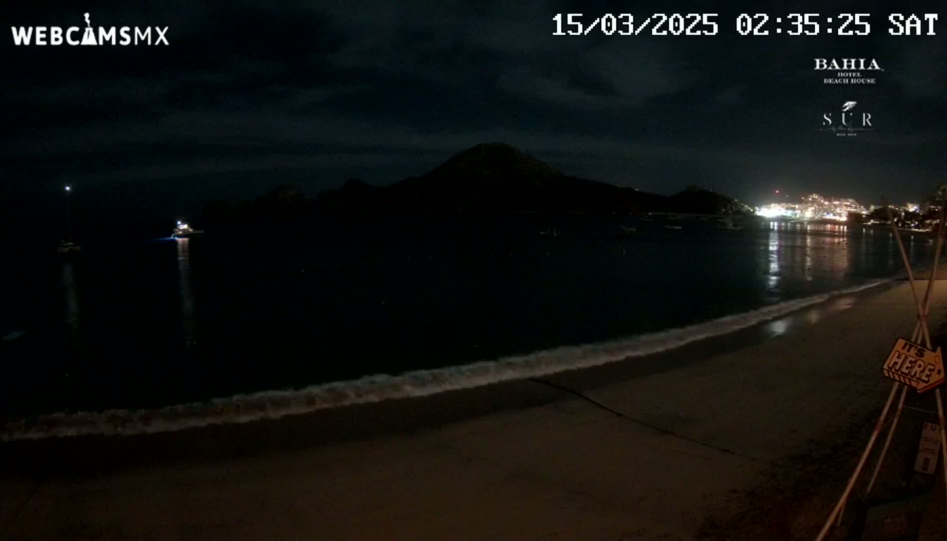 A dark, nighttime beach scene shows a calm ocean with gentle waves lapping the shore, a distant, silhouetted mountain, and a few lights visible across the water.