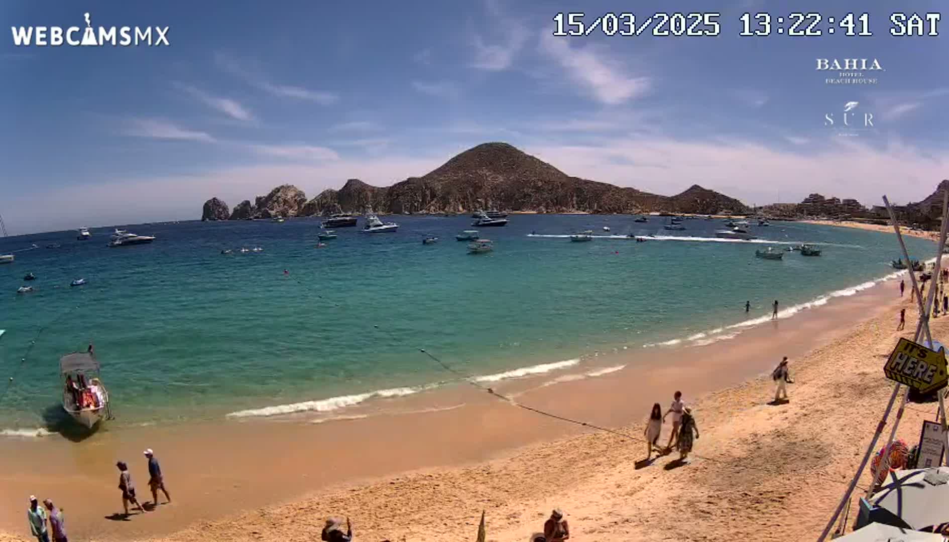 A sandy beach with turquoise water and several boats in the bay under a sunny sky.
