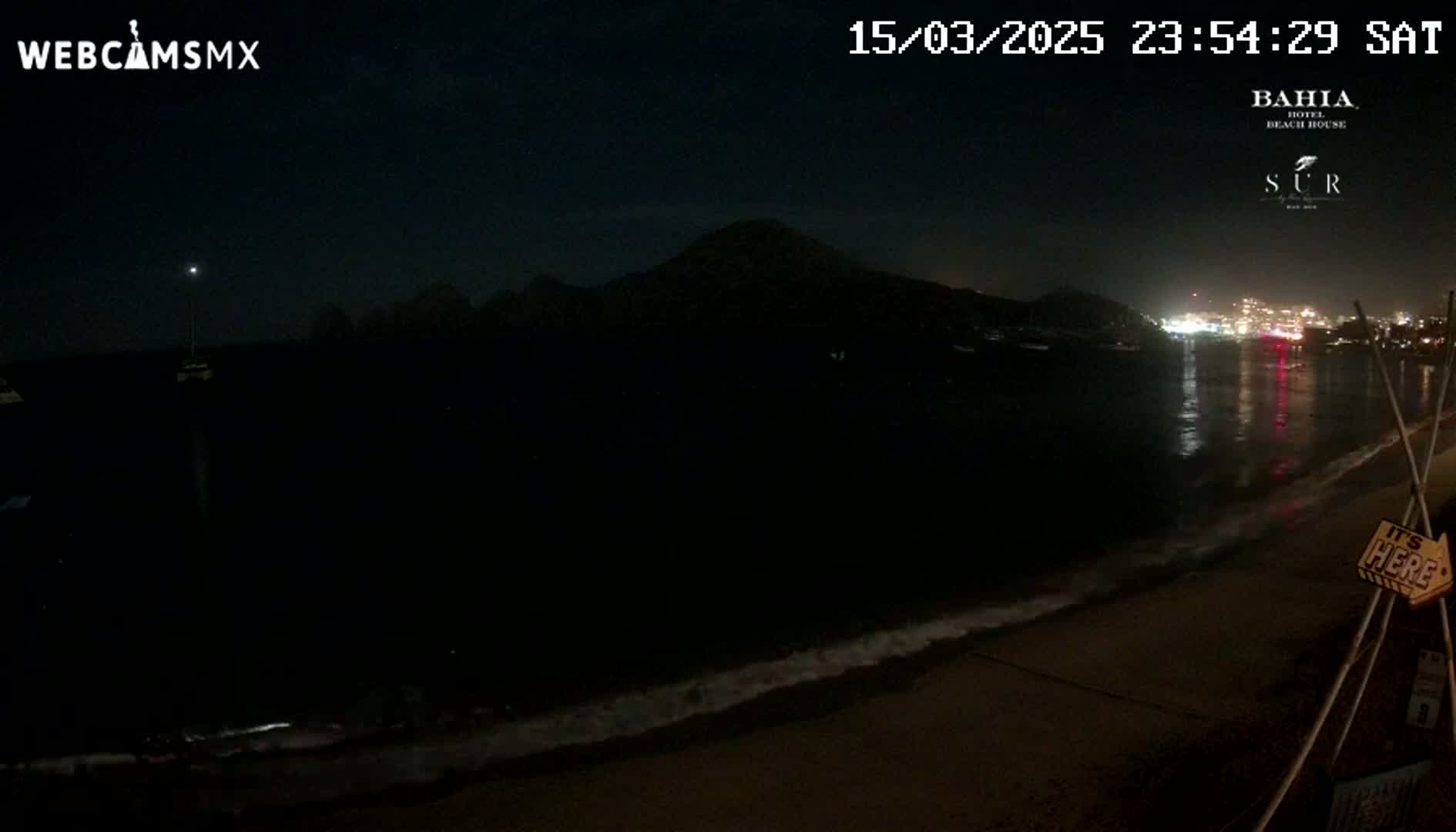 A dark nighttime beach scene shows a calm ocean lapping gently at the shore under a clear, starry sky, with a distant, dimly lit town visible across the water and a hill silhouetted against the night.
