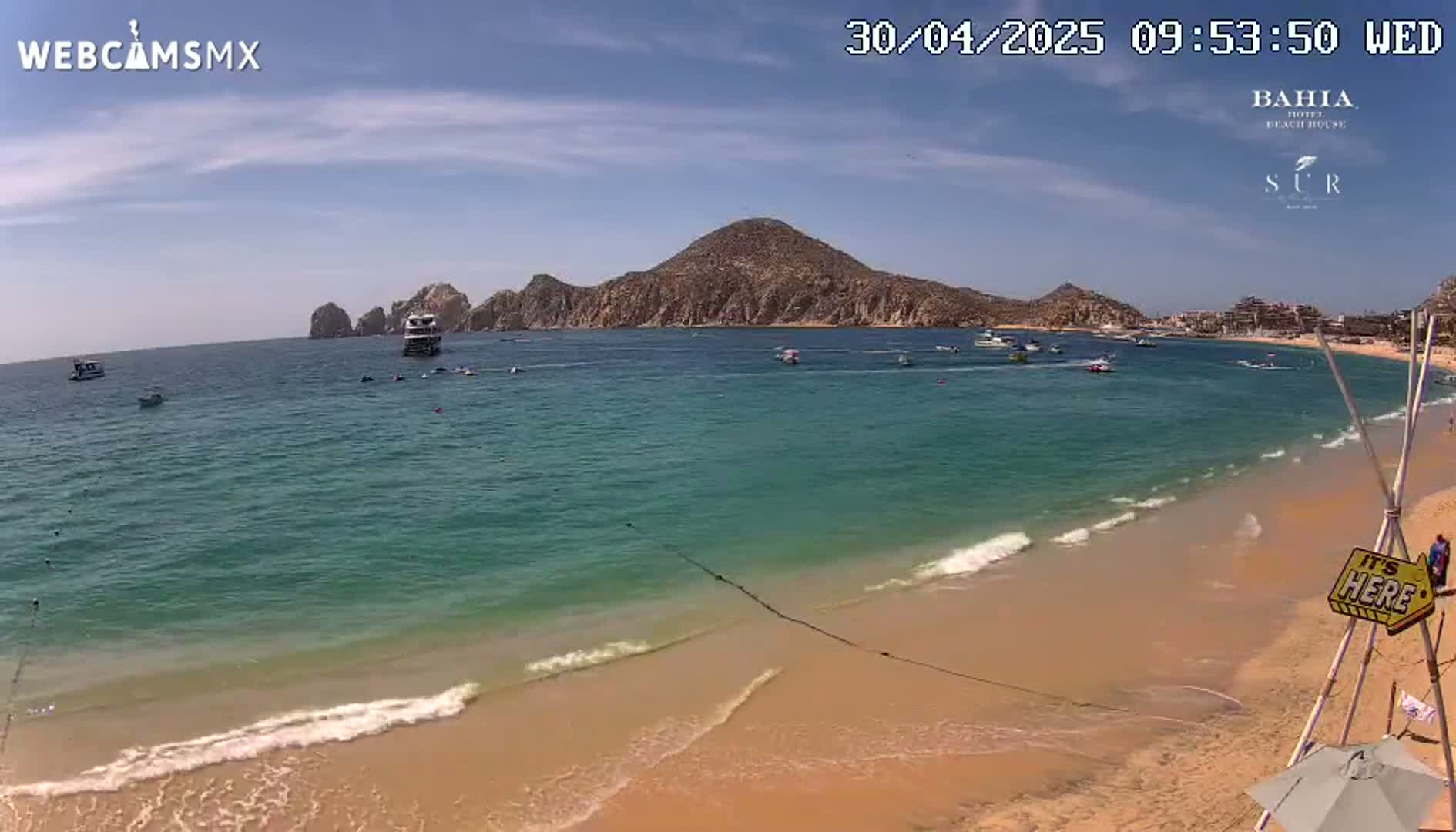 A sandy beach with turquoise water laps gently at the shore, under a sunny sky, with several boats and a large landmass in the distance.