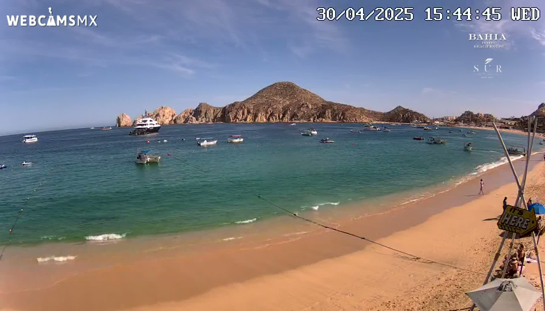 A sandy beach with calm, clear turquoise water, several small boats, a large yacht, and a rocky hill in the background under sunny skies.