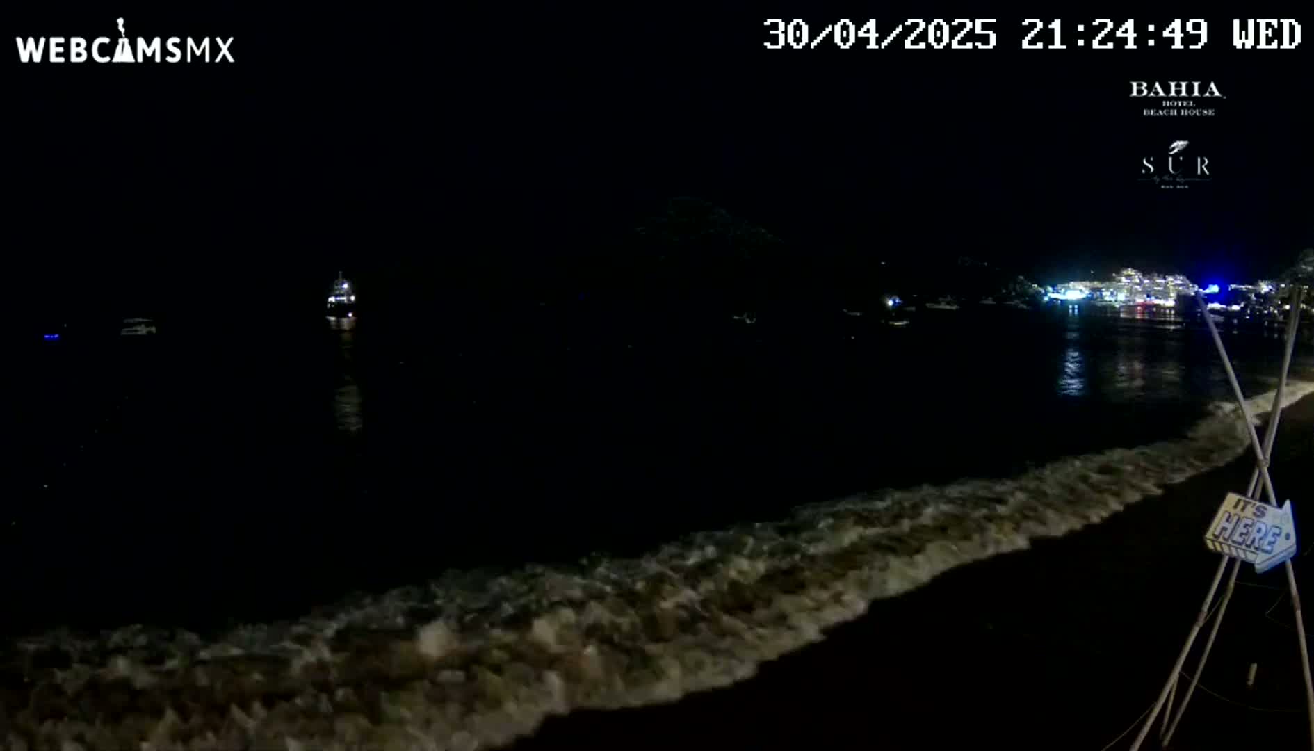 At night, small waves lap a dark beach under a clear, dark sky, with lights visible across the water in the distance.