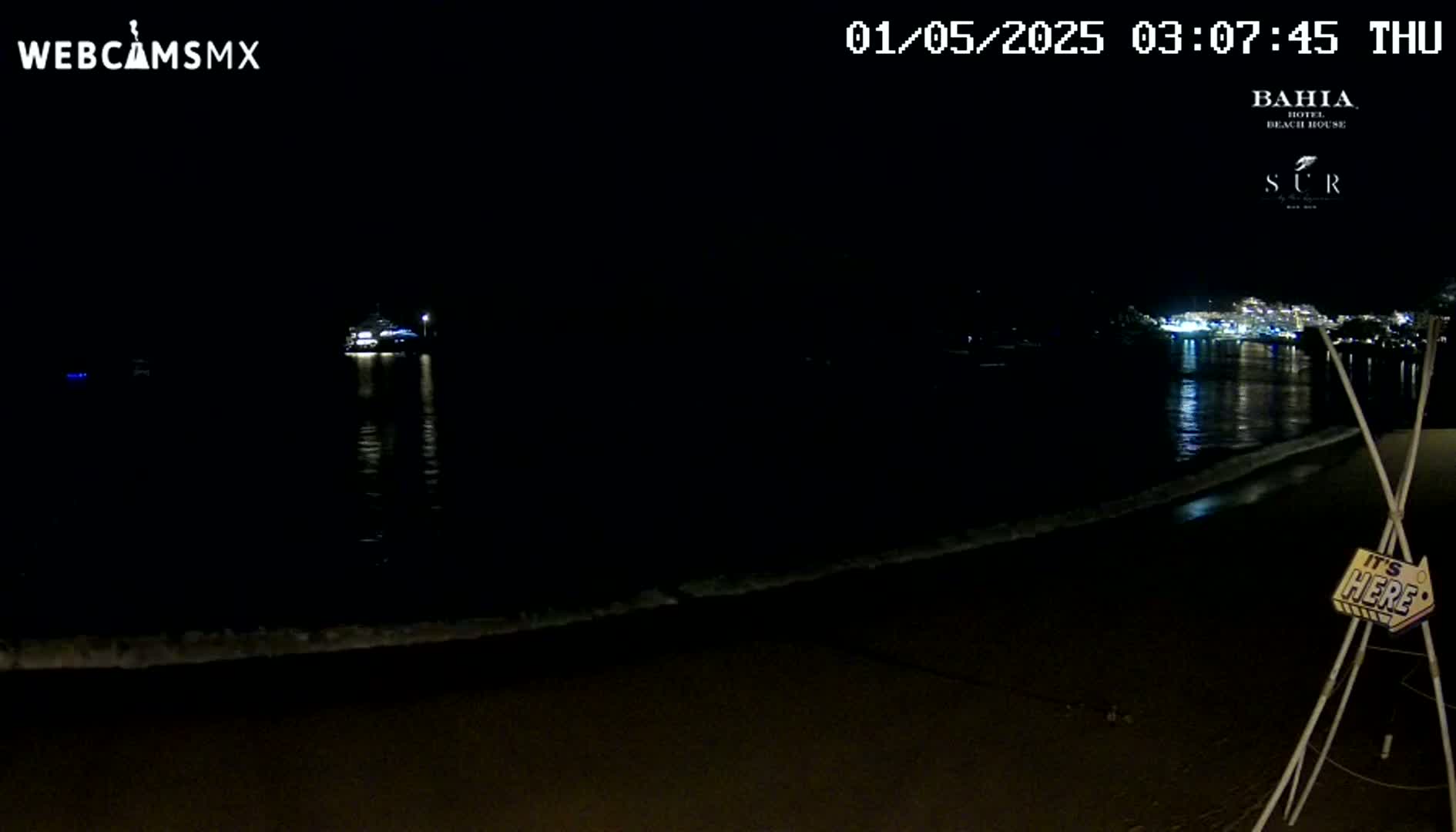 A dark, nighttime beach scene shows gentle waves lapping the shore under a clear, starless sky, with lights visible across the water.
