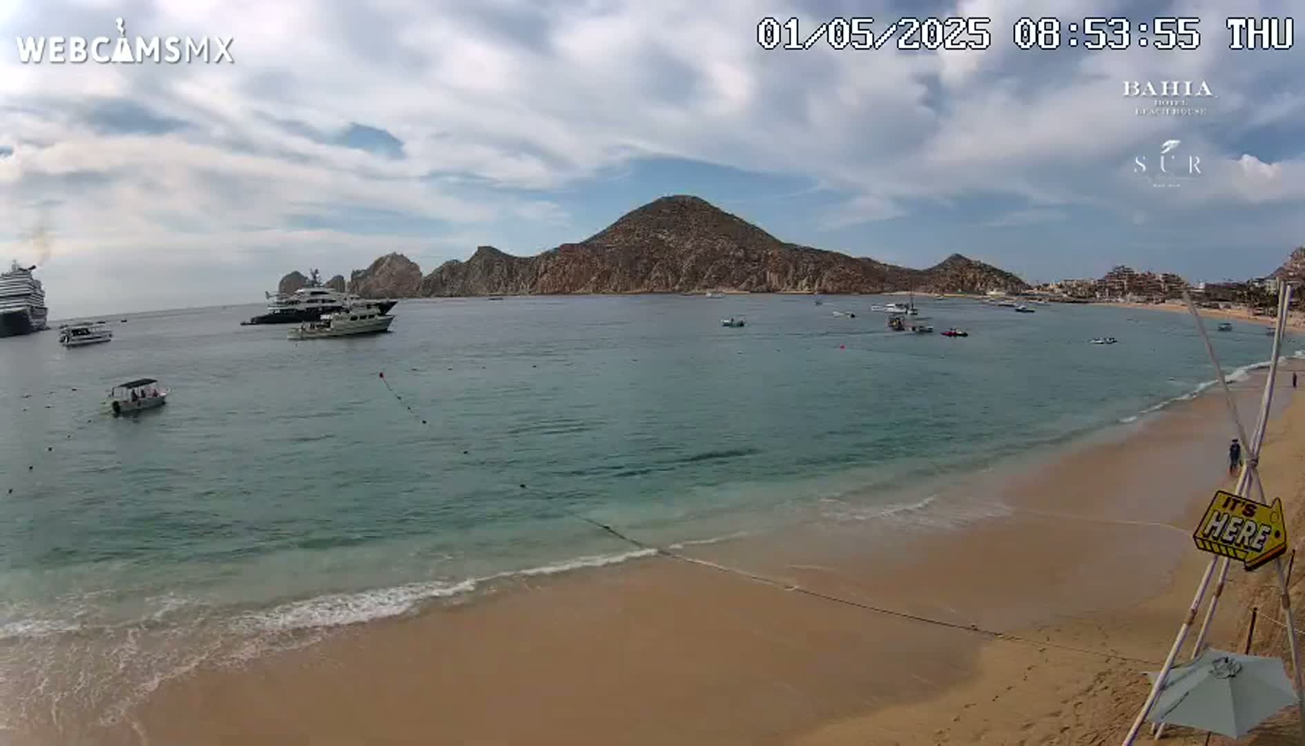 A partly cloudy day shows a sandy beach with calm, turquoise water; several boats and a large cruise ship are in the bay, with a mountainous peninsula in the background.