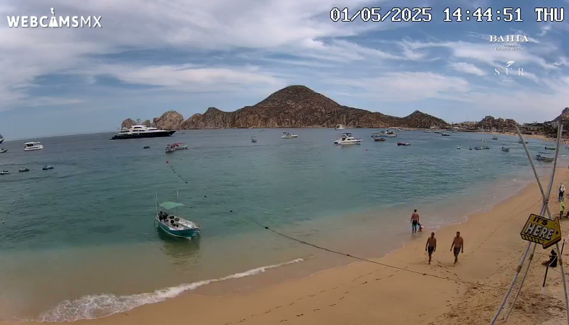 A sandy beach with calm, clear water, several boats, and a few people on the shore under a partly cloudy sky.