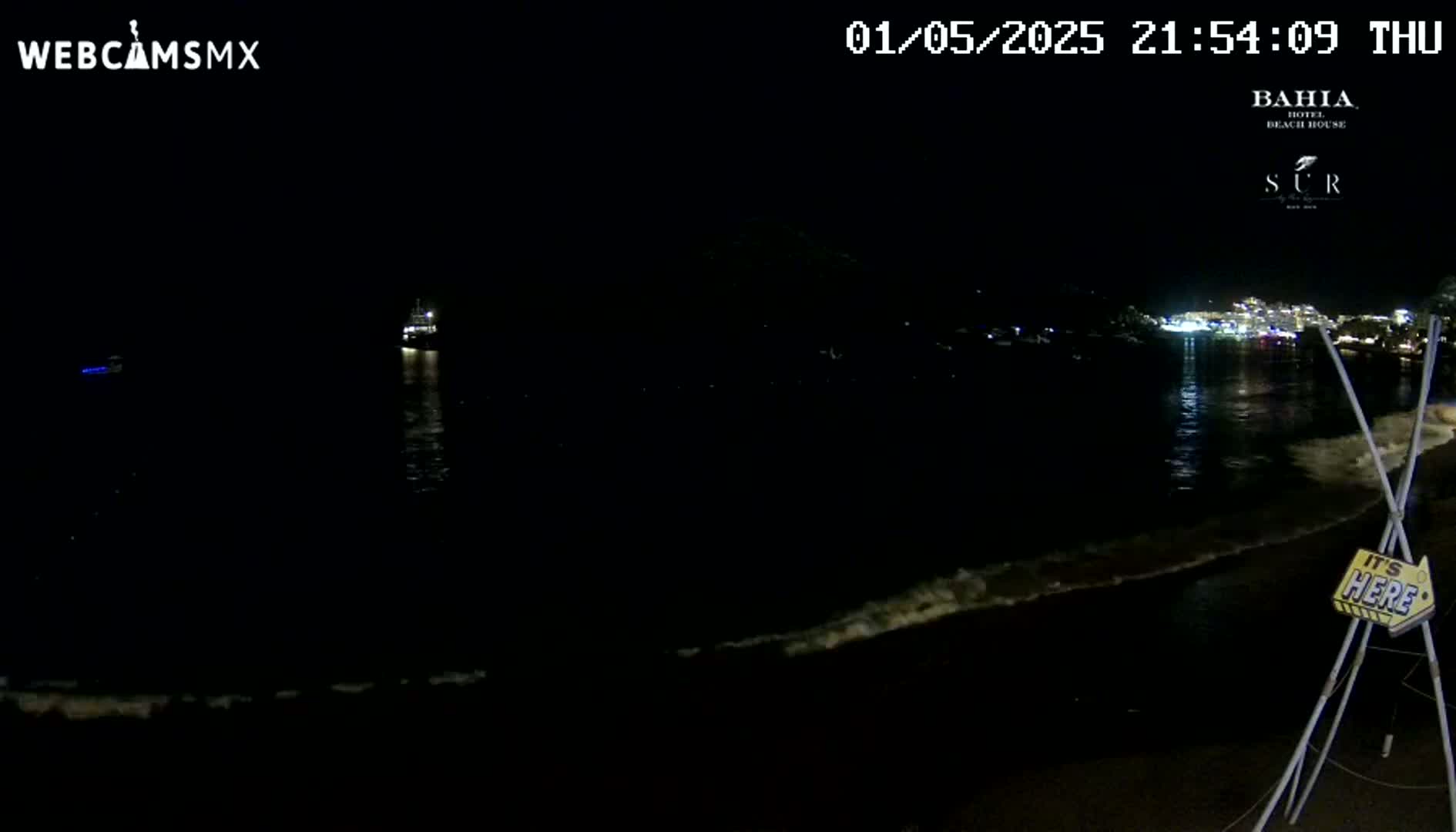 A dark, nighttime beach scene shows gently lapping waves on the shore with a distant, faintly lit town across the water.
