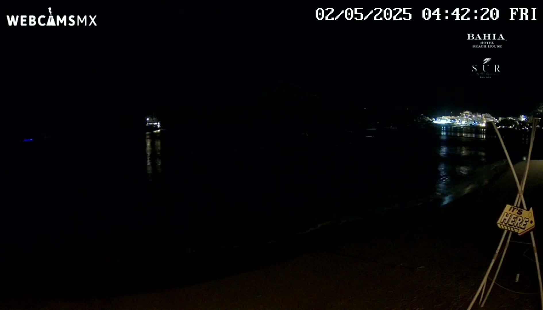 A dark nighttime beach scene shows a calm body of water reflecting distant lights, with a small boat visible in the distance.