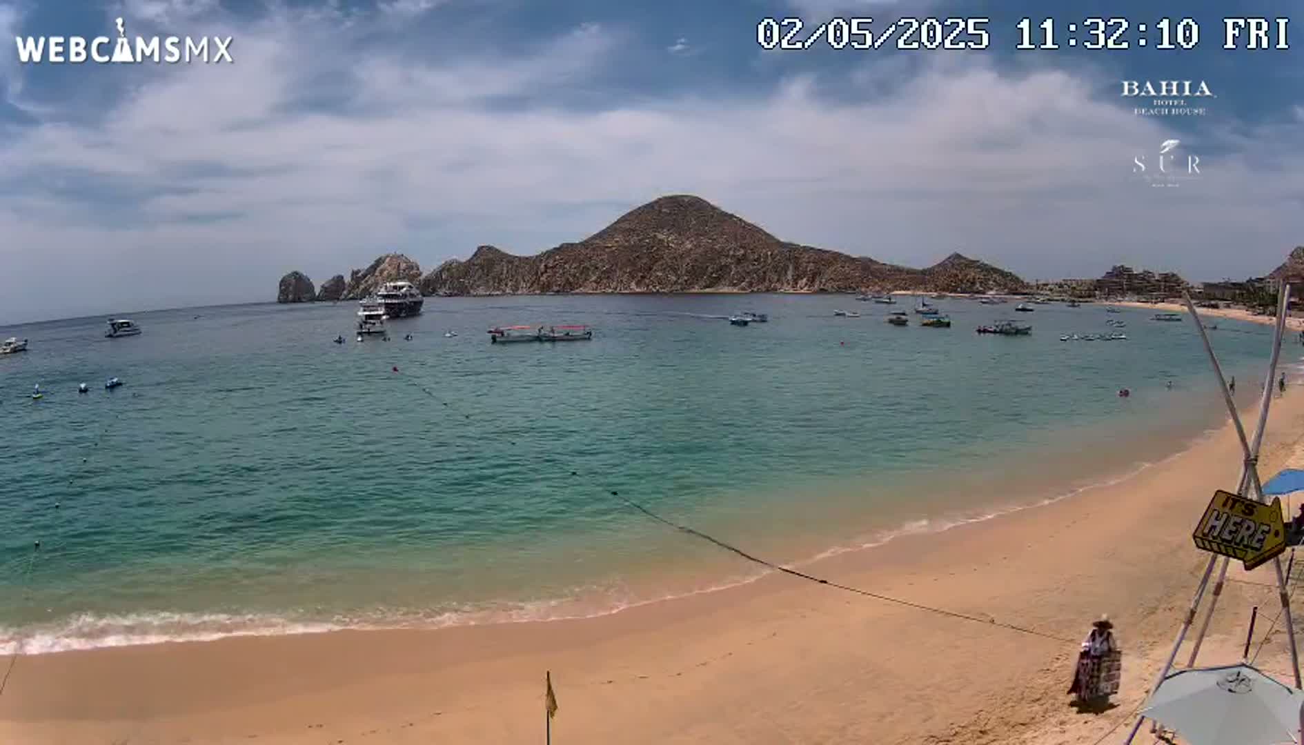 A sandy beach with calm, turquoise water laps gently at the shore, under a partly cloudy sky, with several boats and people visible in the distance.