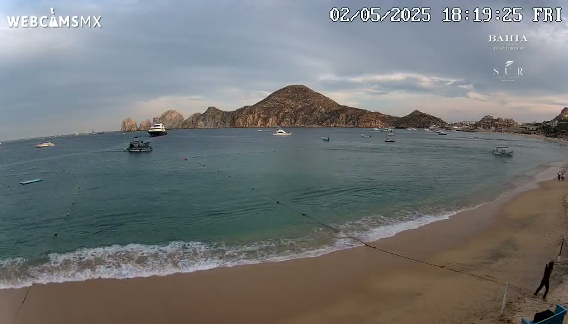 A sandy beach with gentle waves laps the shore, overlooking a calm bay with several boats and a large landmass in the distance under a mostly cloudy sky.