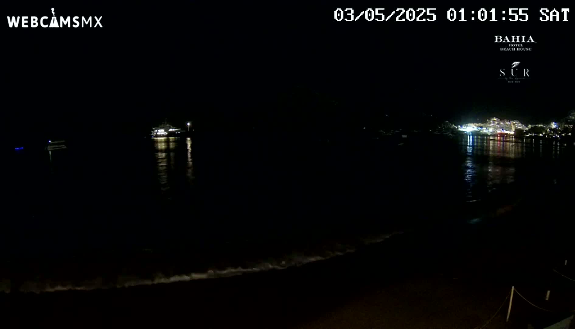 A dark, nighttime beach scene shows calm water reflecting the lights of a town across the bay, with a few boats visible on the water.