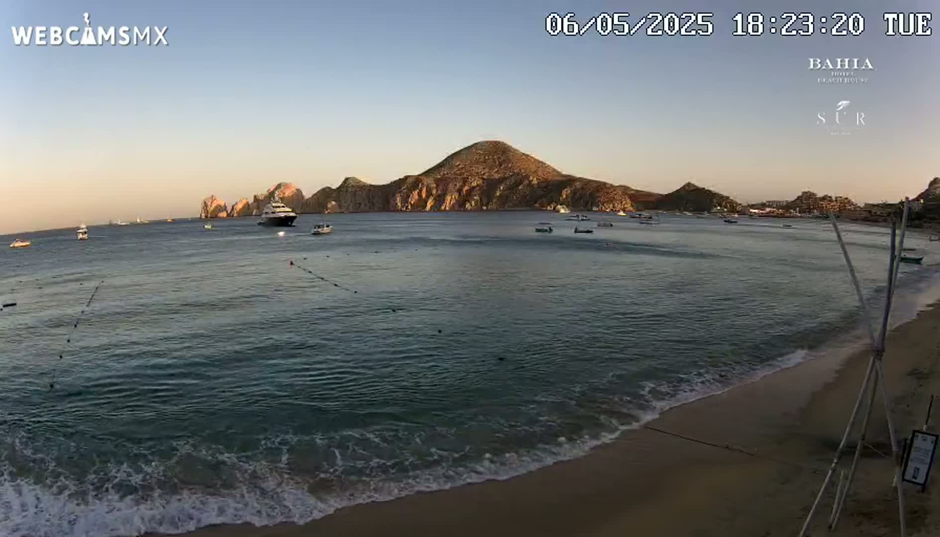 A calm ocean with gentle waves laps a sandy beach under a clear, twilight sky, with several boats visible on the water and hills in the distance.