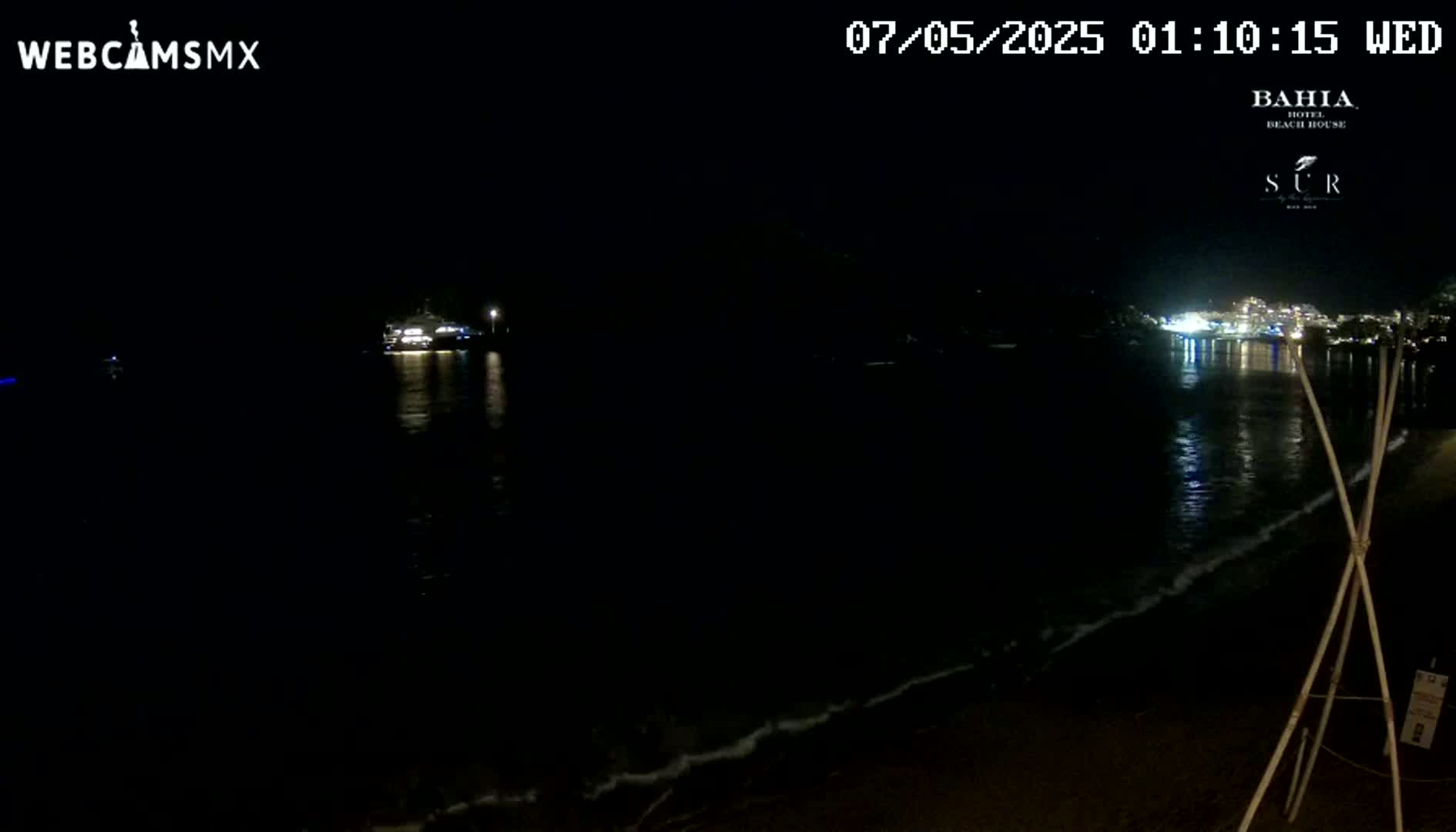 A dark night scene shows a calm body of water lapping at a sandy shore, with lights visible across the water and in the distance.