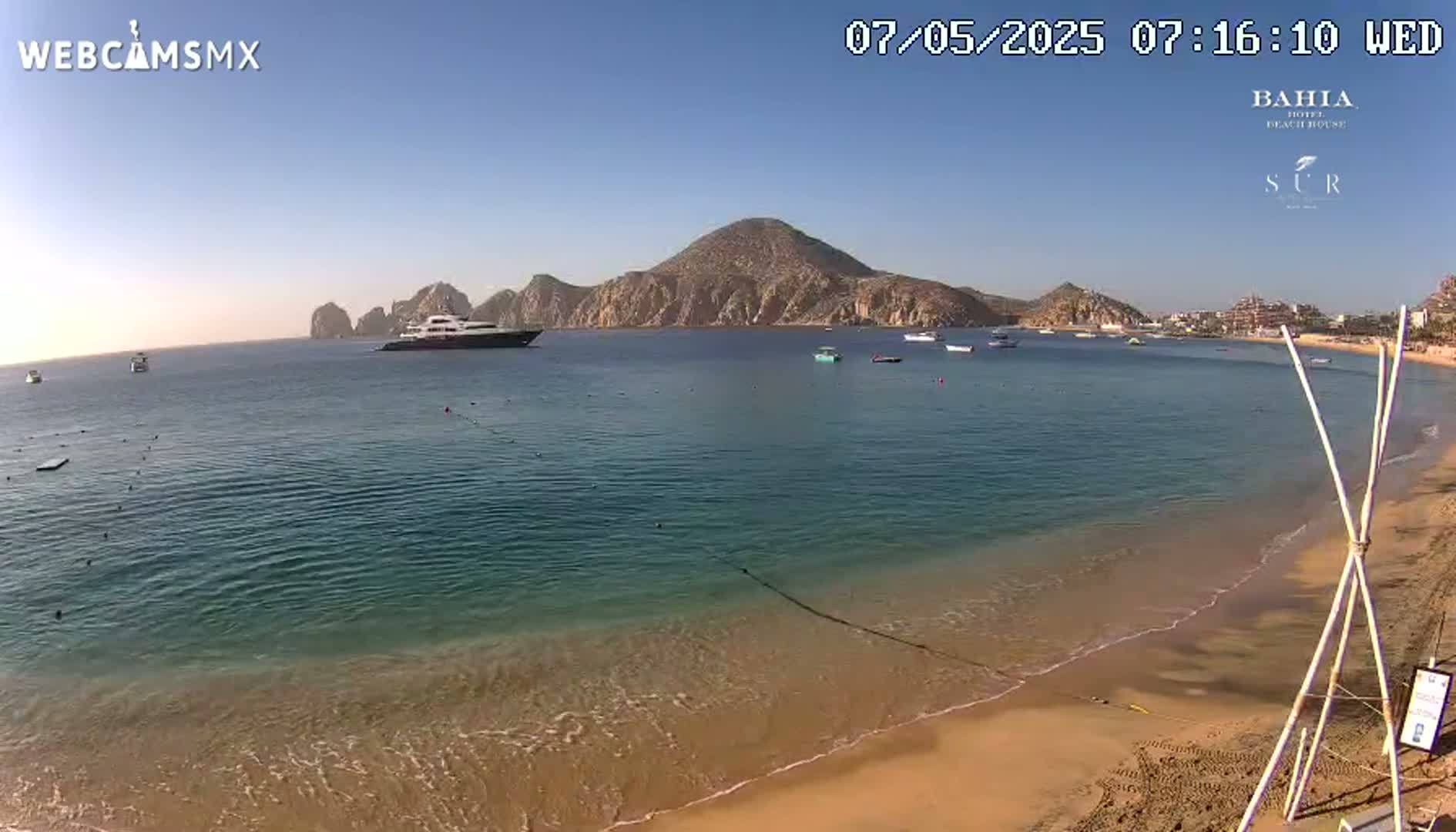 A sandy beach with calm, clear turquoise water, a large yacht, and a mountainous backdrop under a clear, sunny sky.