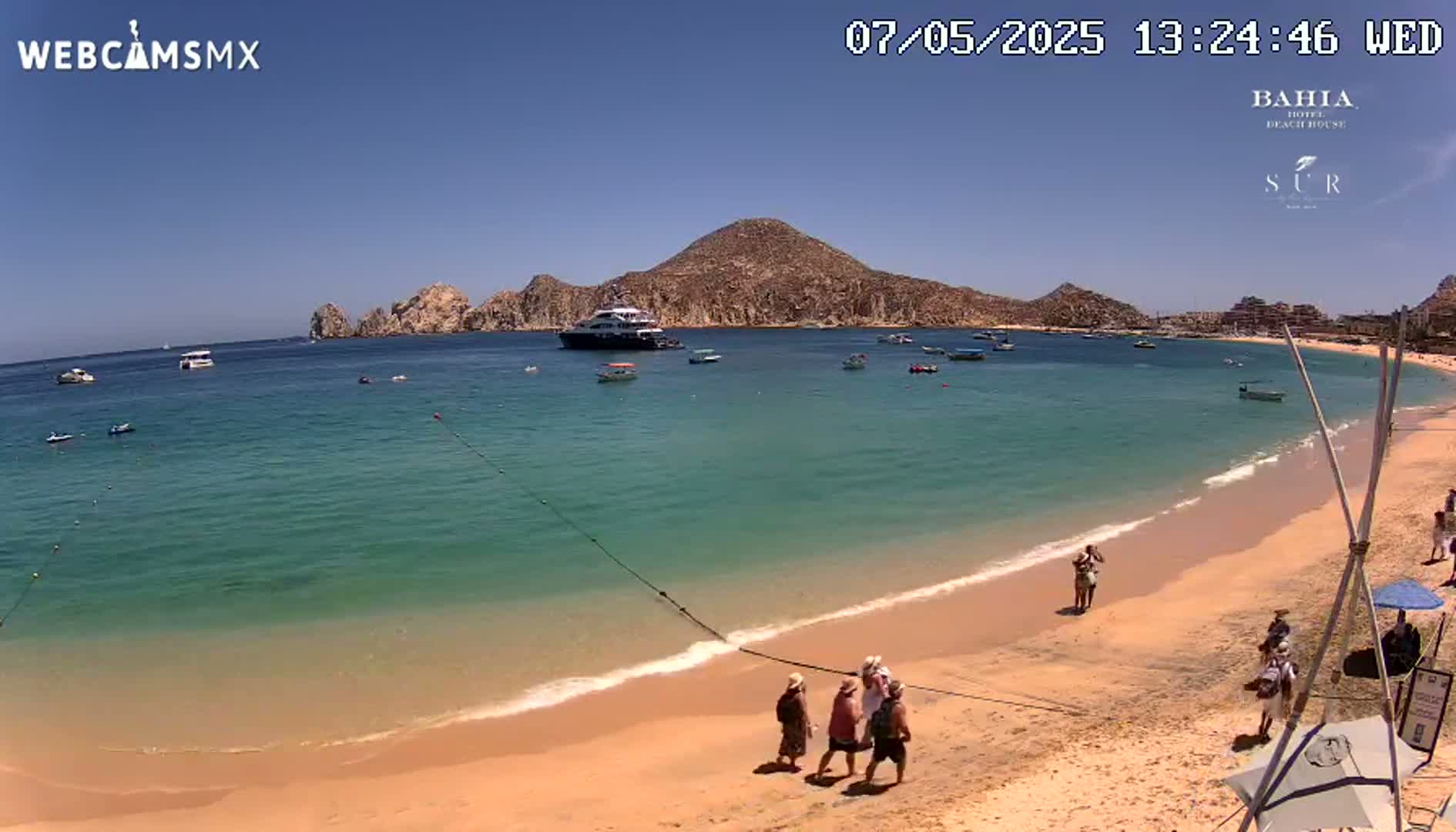 A sandy beach with turquoise water, several boats, and a few people walking along the shore under sunny skies.