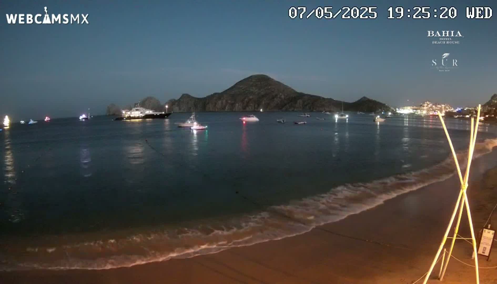 A sandy beach at night, under a twilight sky, shows gentle waves lapping the shore, with several boats and a large yacht visible in the calm water, and a dark mountain in the background.