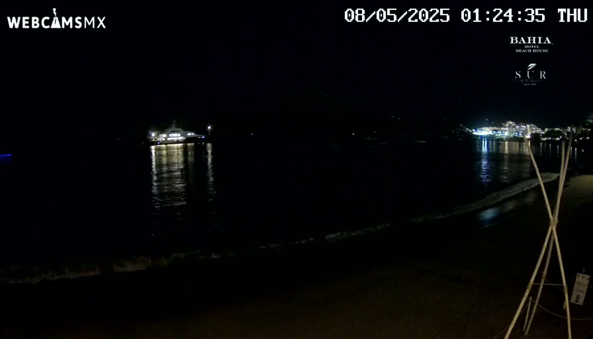 A nighttime beach scene shows a calm ocean with gentle waves lapping the shore, a boat in the water, and illuminated buildings across the bay.