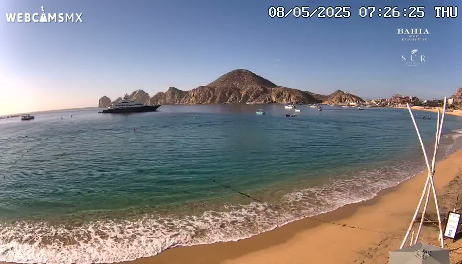 A sandy beach with calm, clear turquoise water laps gently at the shore under a clear blue sky, with a large yacht and several smaller boats visible in the bay beyond a rocky, mountainous coastline.