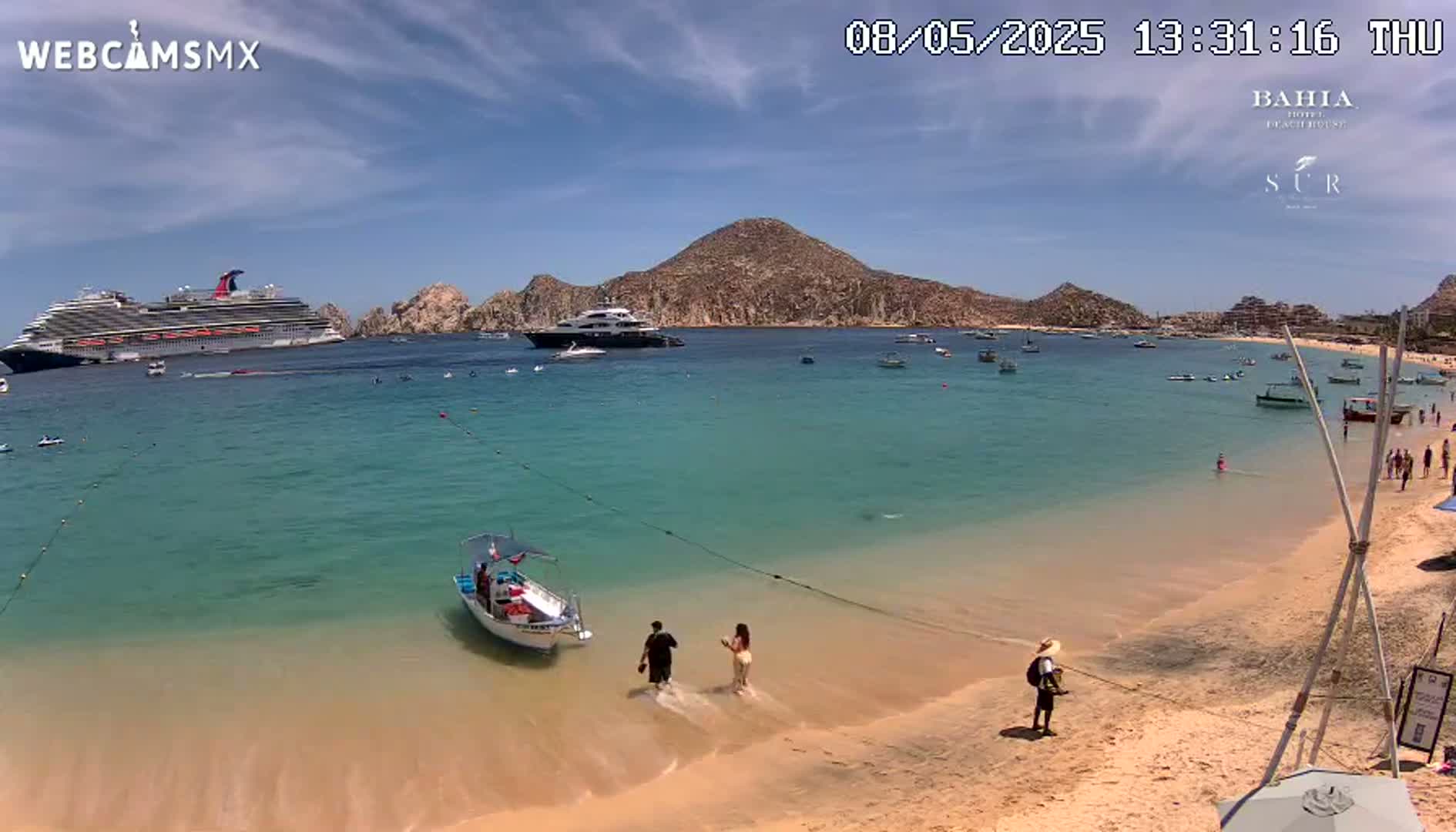 A sandy beach with turquoise water shows a large cruise ship and a yacht in the distance under a mostly sunny sky; several small boats and people are visible near the shore.