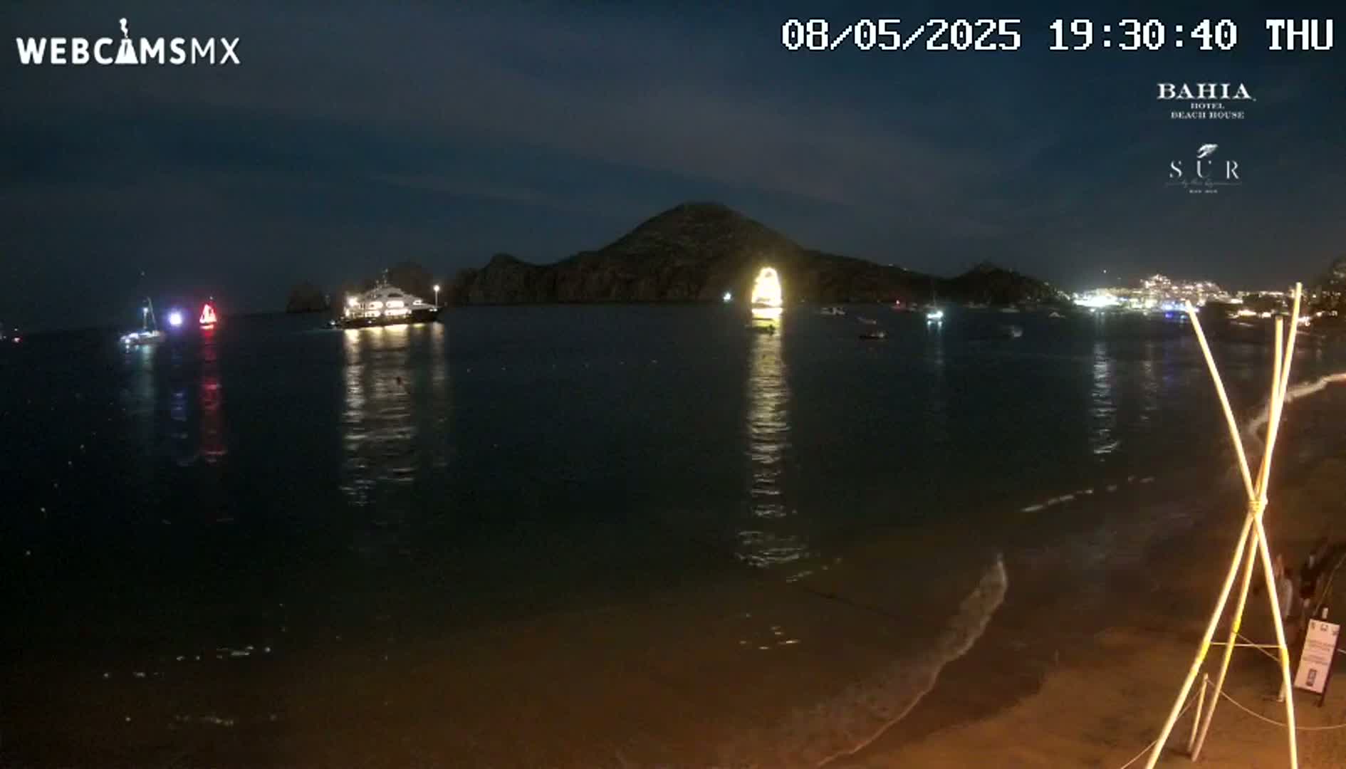 A nighttime view of a sandy beach with calm waves, several boats illuminated on the dark water, and a brightly lit hill in the background.