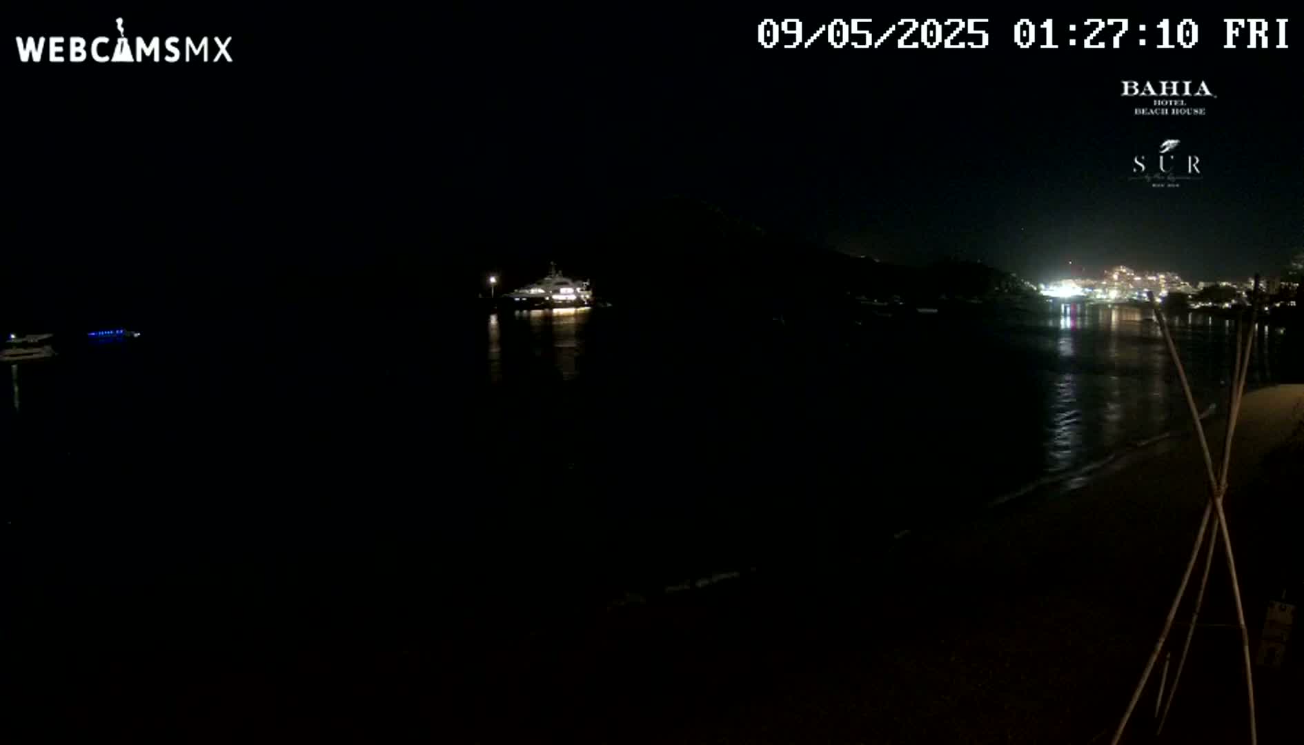 A dark nighttime beach scene shows a body of water reflecting distant lights, with a large boat visible in the middle distance.
