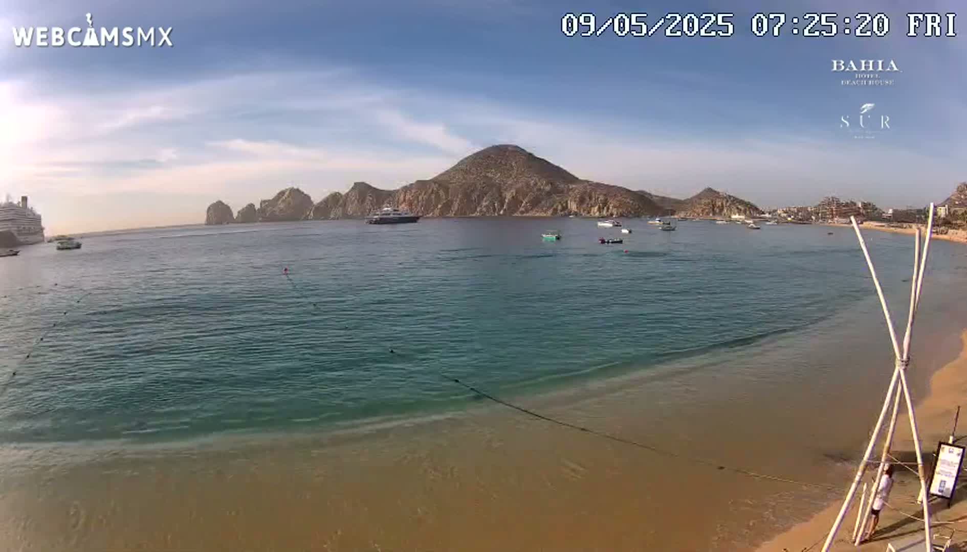 A sandy beach with calm, clear turquoise water, a large cruise ship and several smaller boats in the distance, under a mostly sunny sky.