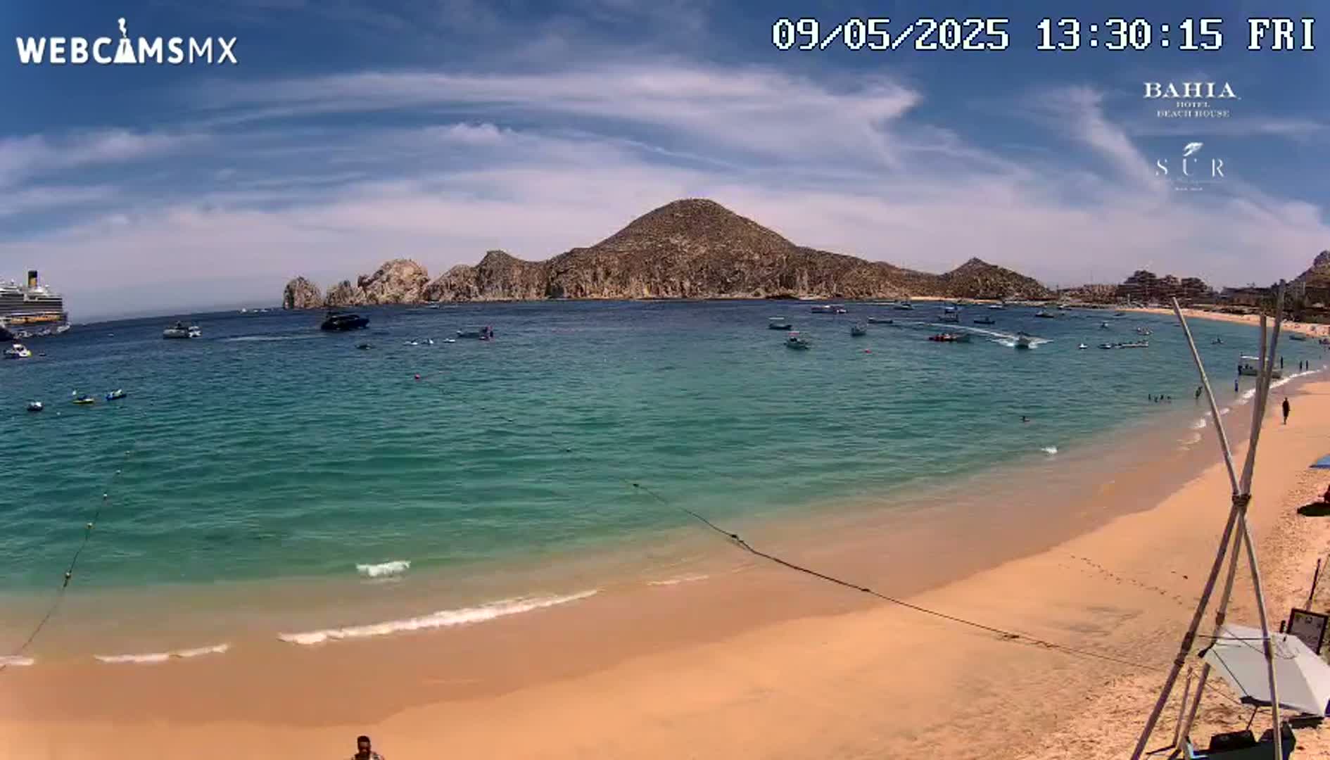 A sandy beach with turquoise water and a large rocky landmass in the background under a mostly sunny sky.