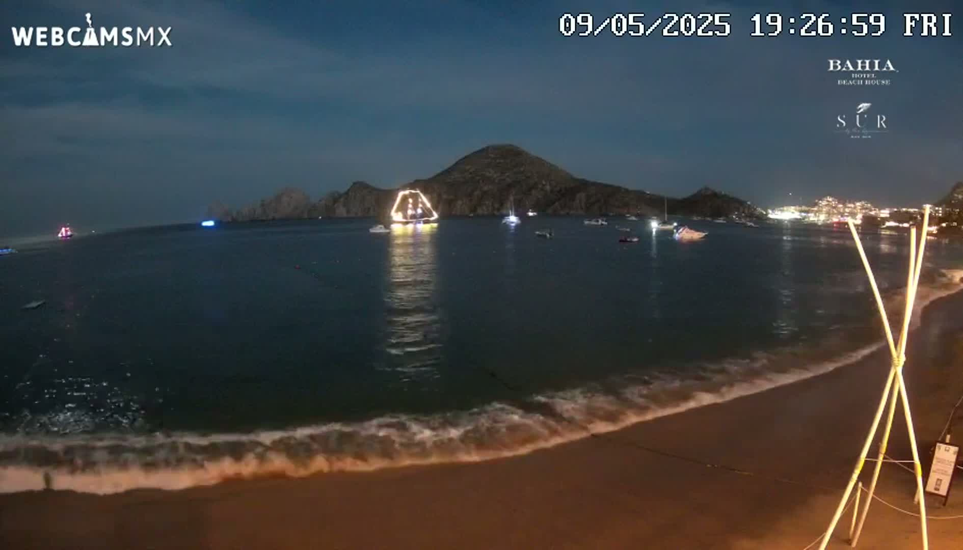 A nighttime beach scene shows a calm ocean with small waves lapping the shore, several boats on the water, and a large, illuminated structure in the distance near a dark mountain under a twilight sky.