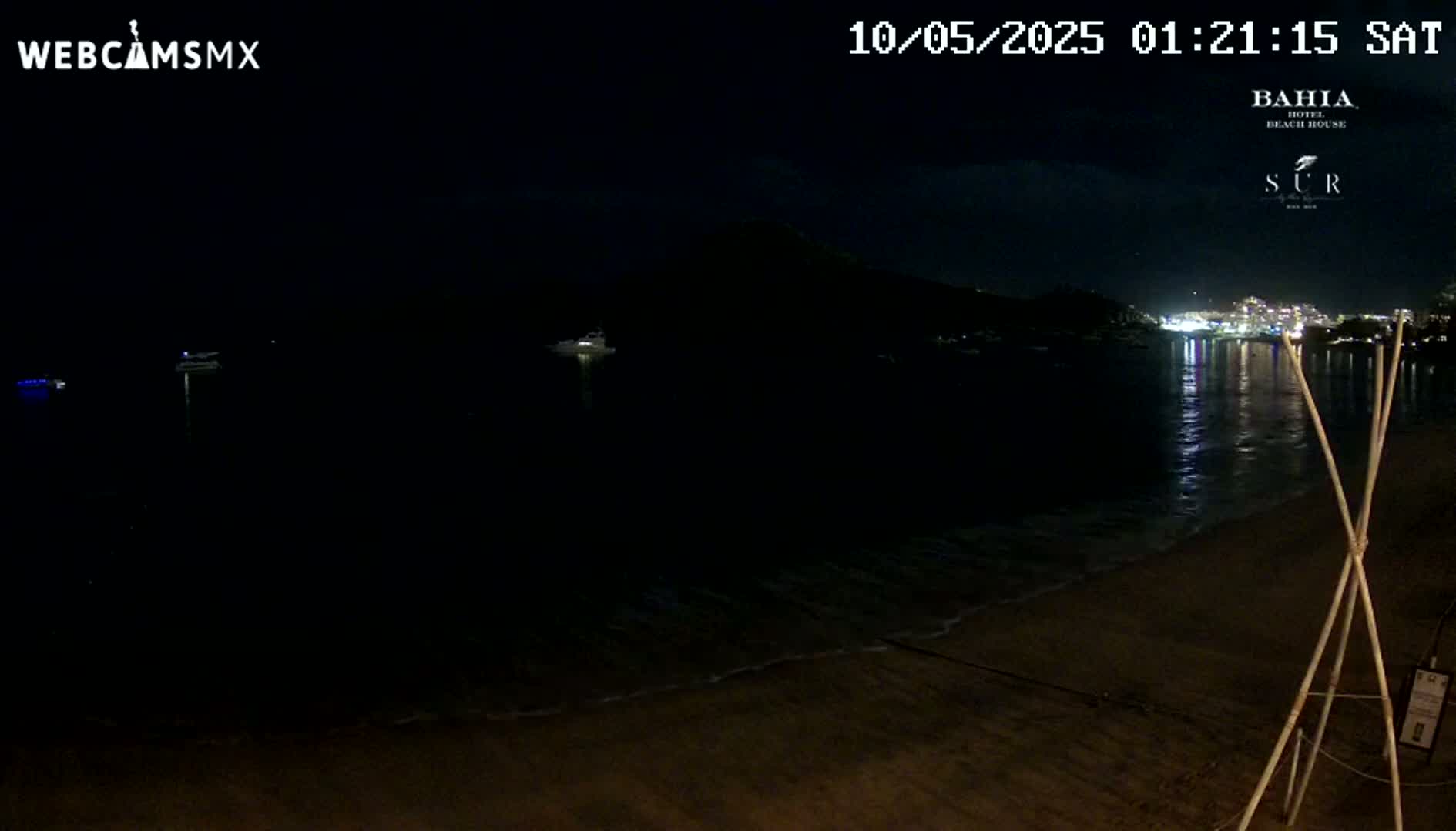 A dark, nighttime beach scene shows a calm, dark ocean reflecting a distant town's lights, with a sandy shore in the foreground.