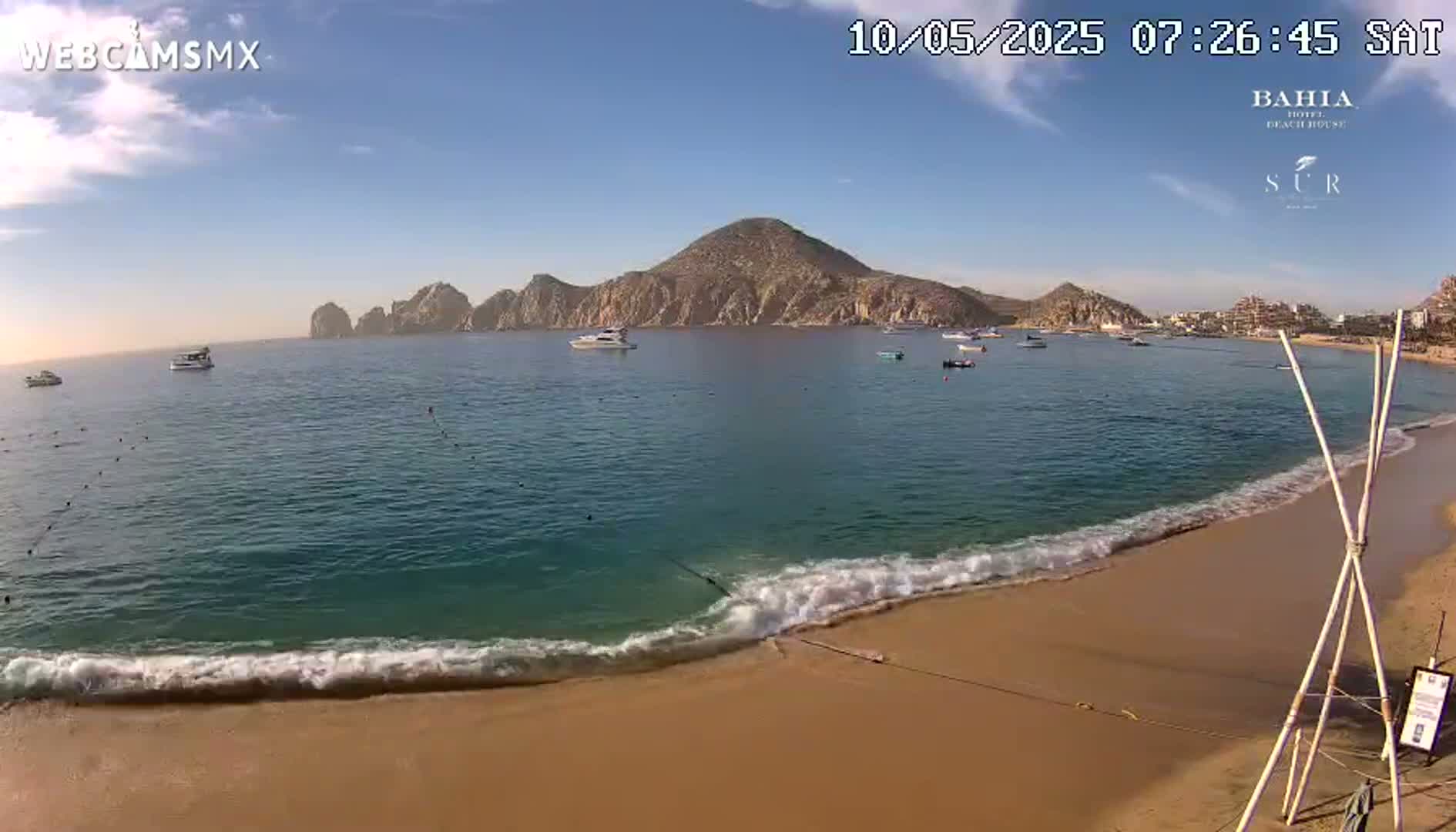 A sandy beach with gentle waves laps the shore, showcasing a clear blue ocean with several boats and a mountainous peninsula under a sunny sky.